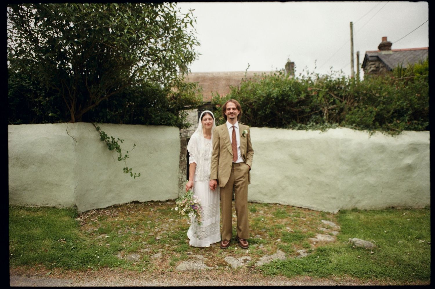 Bride and groom posing together in front of a white stone wall in a garden on their wedding day.