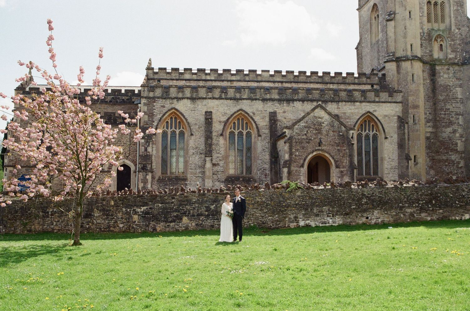Bride and groom standing together on a green lawn beside a historic stone church with pink blossom tree.