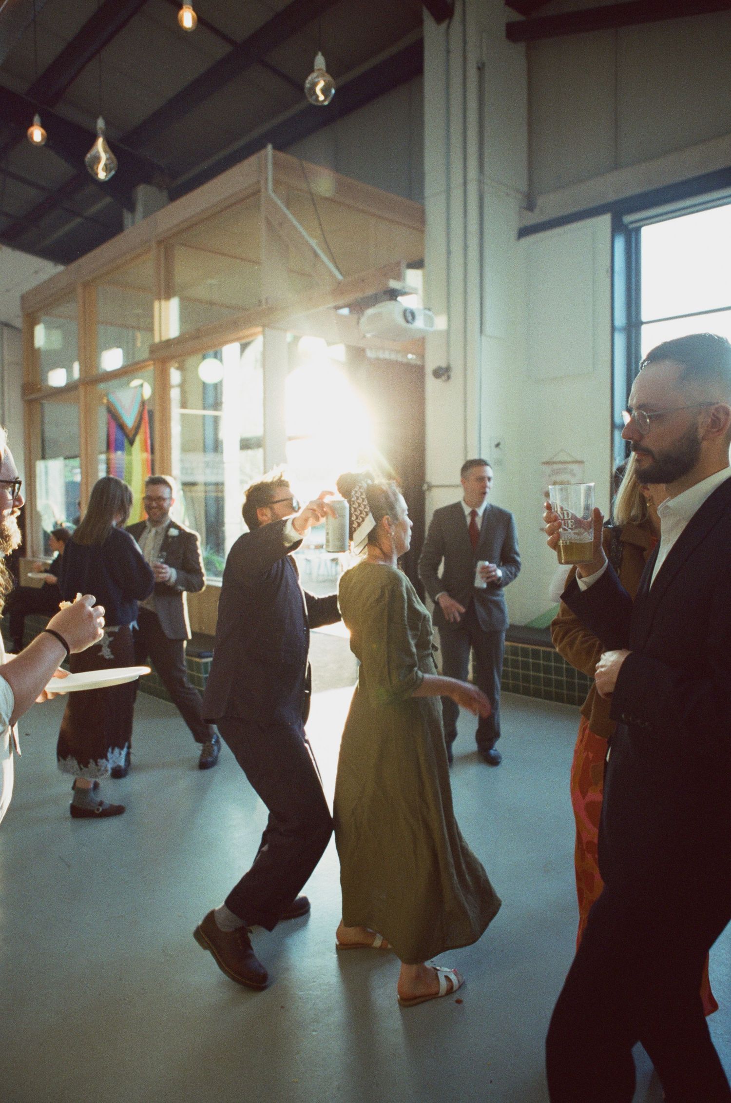 Wedding guests dance and mingle in a sunlit industrial venue with exposed ceiling lights and green tile walls.