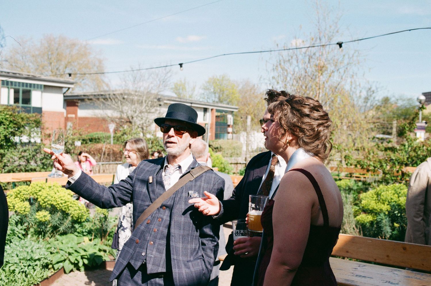 Two elegantly dressed guests chat outdoors at a garden party, one wearing a bowler hat and suit, holding drinks on a sunny day.
