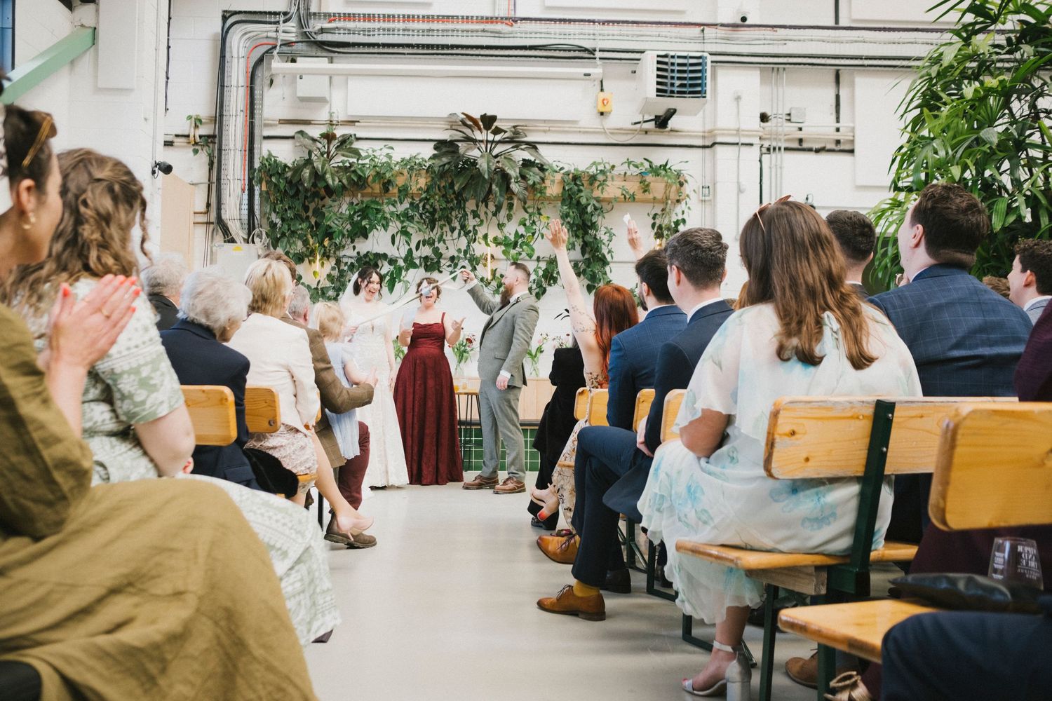Guests seated on wooden benches at an indoor wedding ceremony with lush greenery decorations and a couple at the altar.