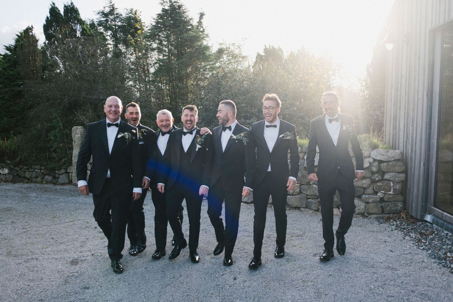 Six groomsmen in black tuxedos walk together outdoors in golden evening sunlight.