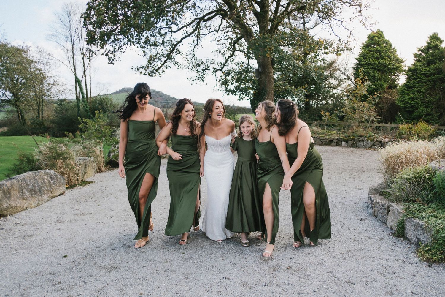 Bride and four bridesmaids in olive green dresses laugh together on a garden pathway.