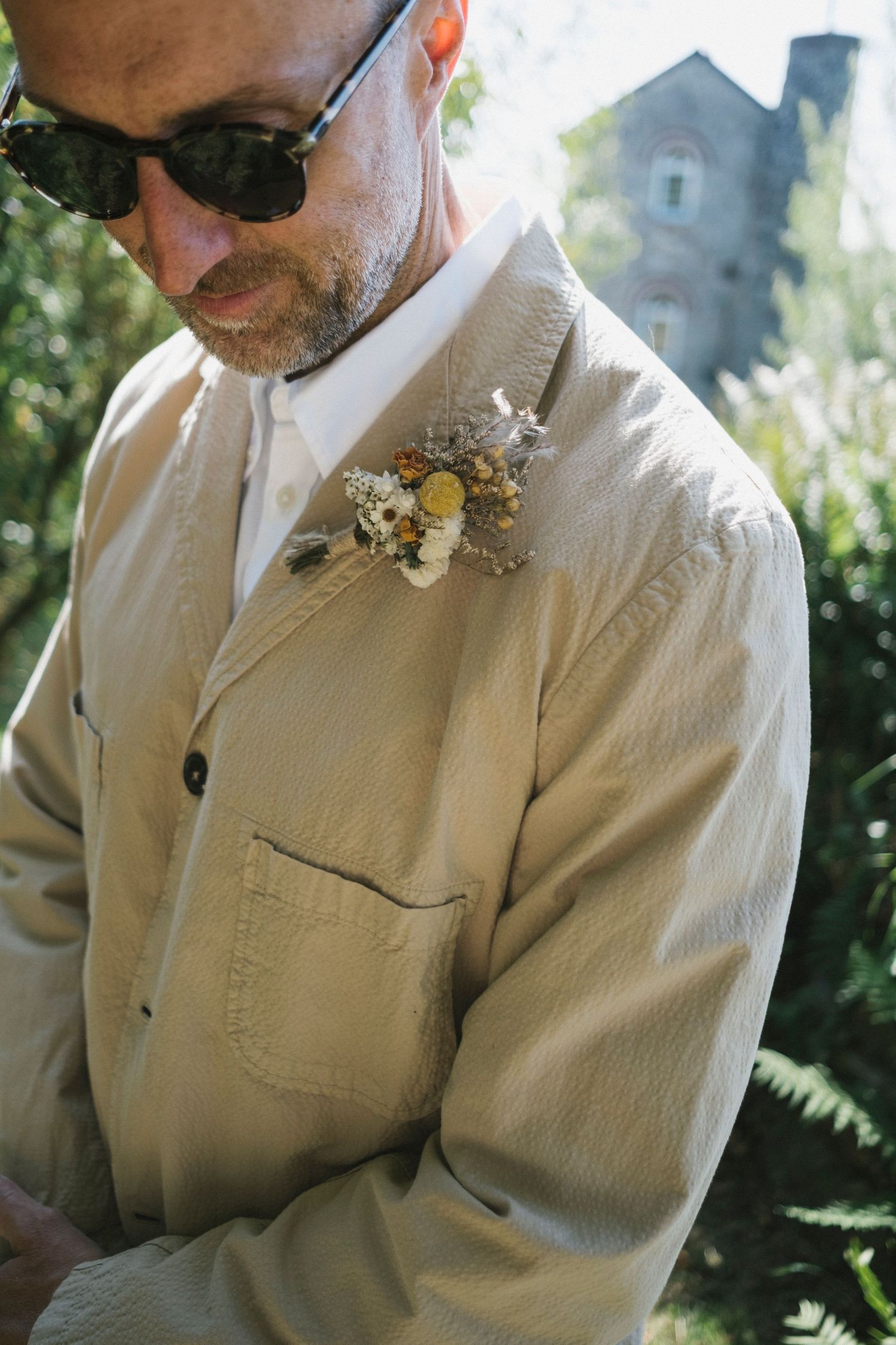 Man in tan suit jacket wearing sunglasses with a rustic boutonniere pinned to his lapel at an outdoor wedding.