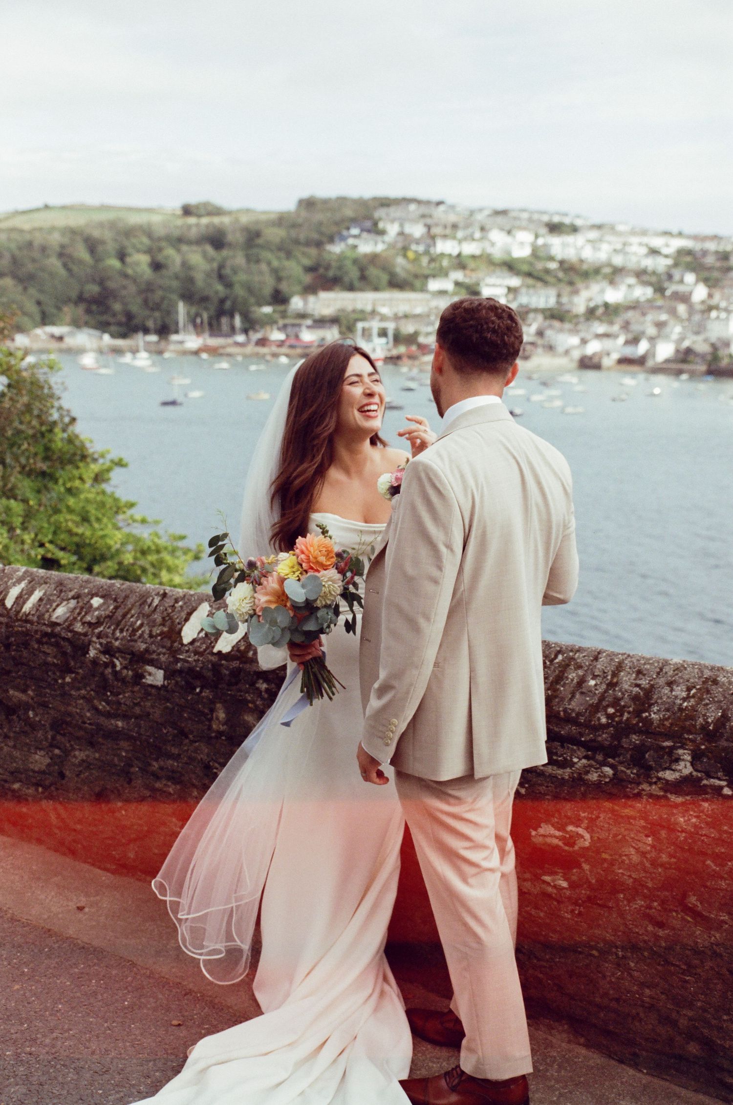 Bride and groom laughing together on a terrace overlooking a coastal town, bride holding a colorful bouquet in a white gown.