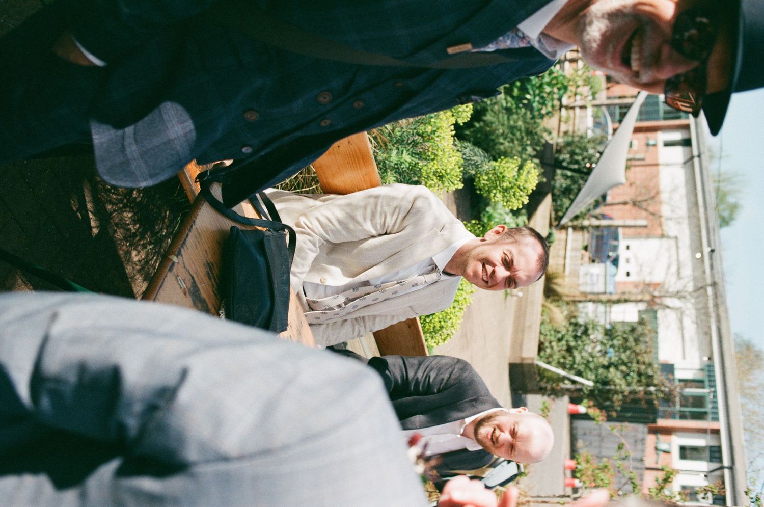 People gathered outdoors at a social event, smiling and conversing near a building with greenery in the background.