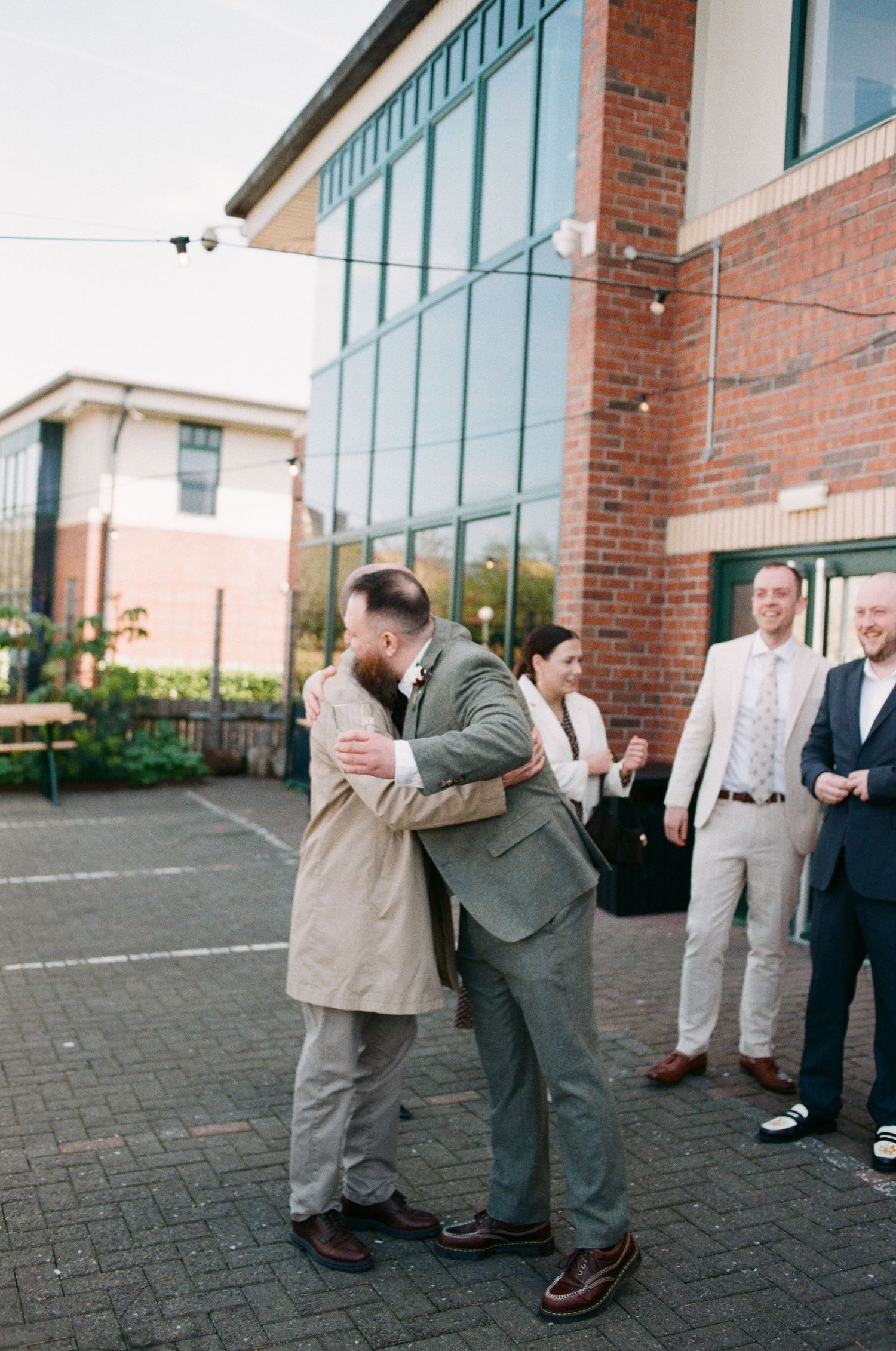 Two men embrace in a heartfelt hug outside a brick building while wedding guests look on smiling.