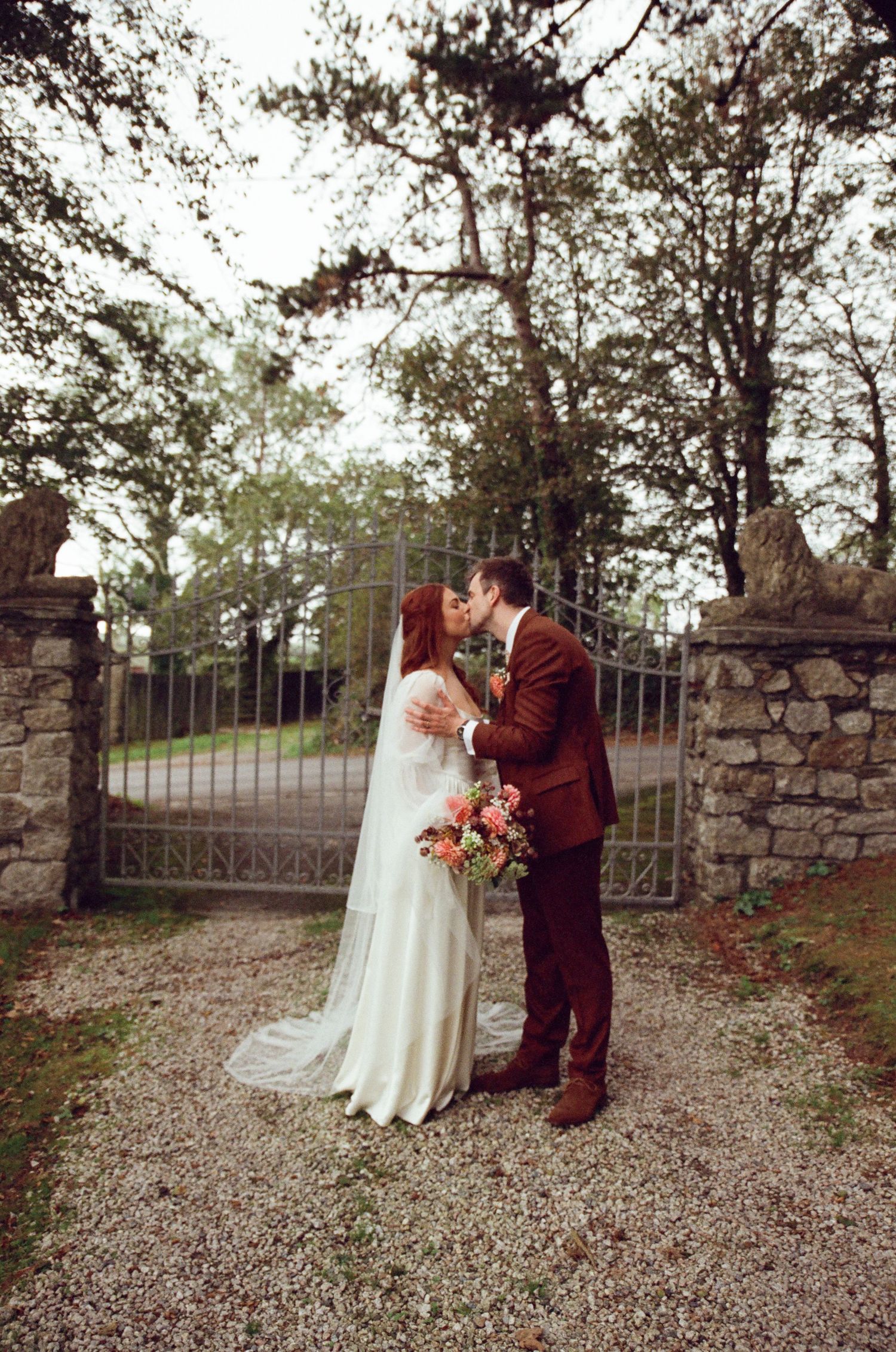 Bride and groom share a kiss at ornate iron gates, she holds a colorful bouquet in a white gown, he wears a brown suit.