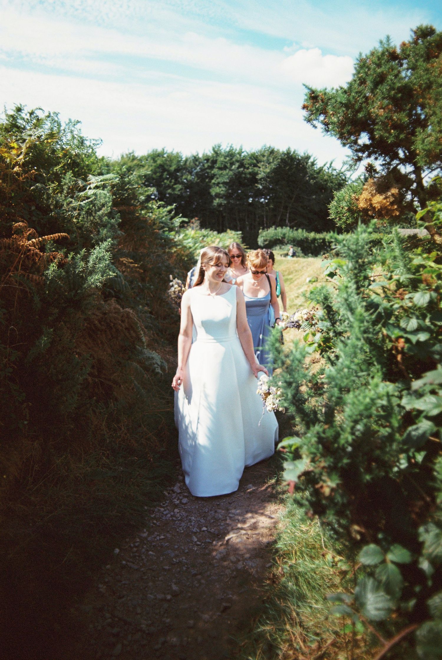 Bride in white gown walking along a narrow garden path with bridesmaids following.