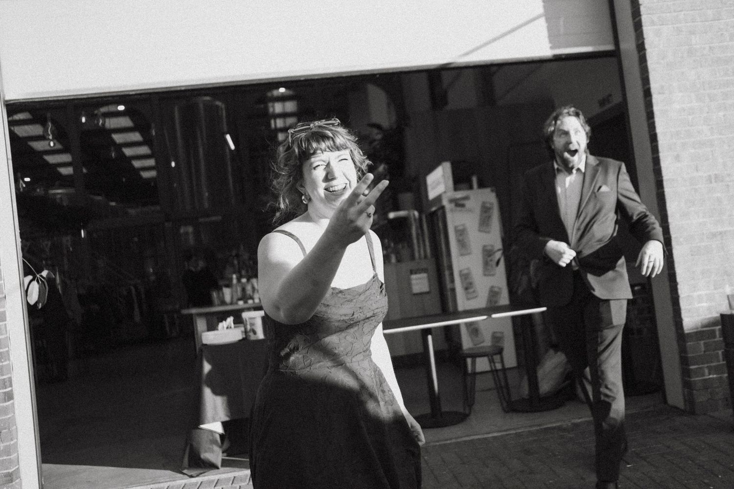 Black and white photo of a woman waving and a man laughing outdoors at a social event.