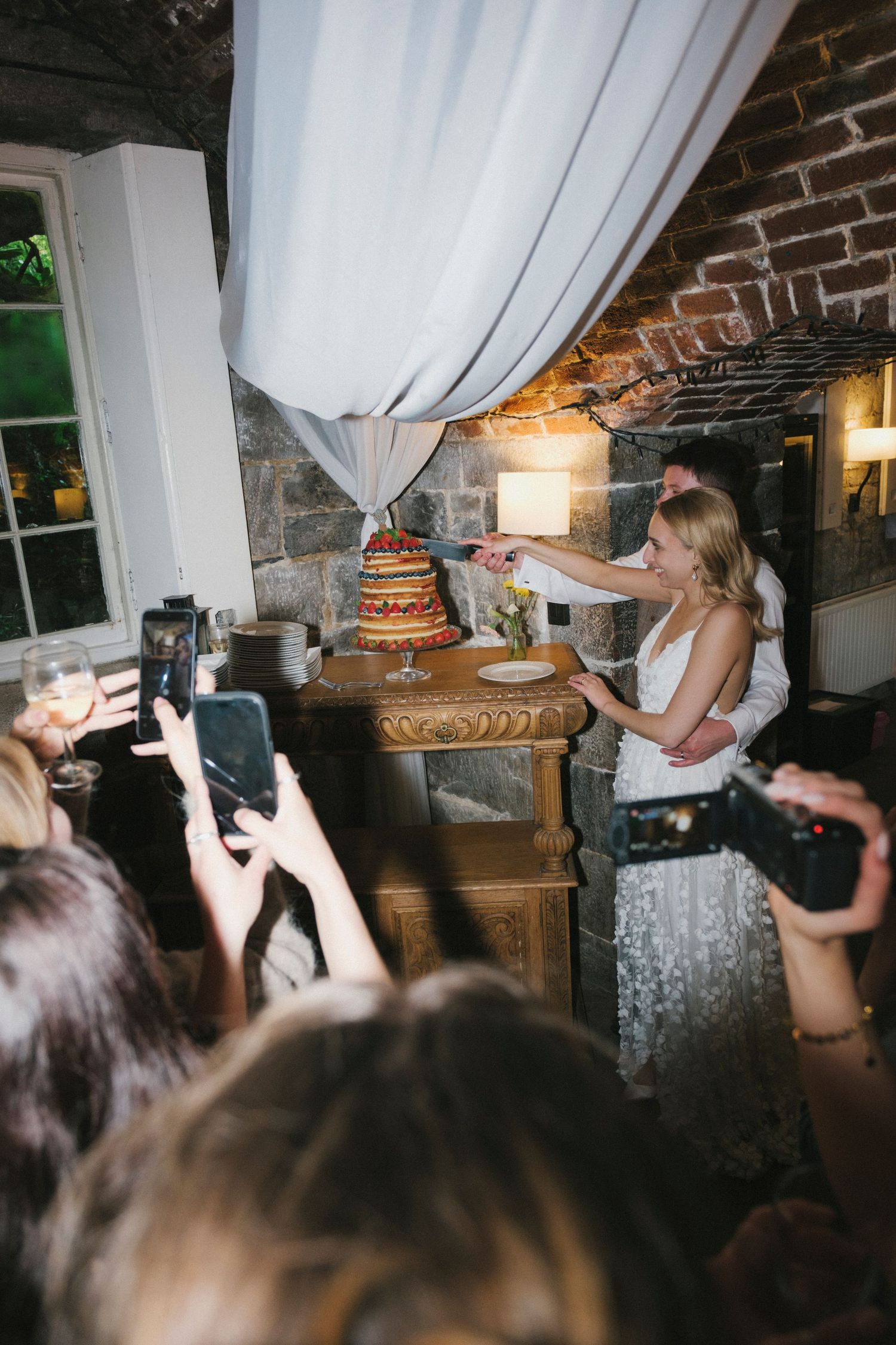 Bride and groom cut their naked layered wedding cake surrounded by guests capturing the moment on phones.