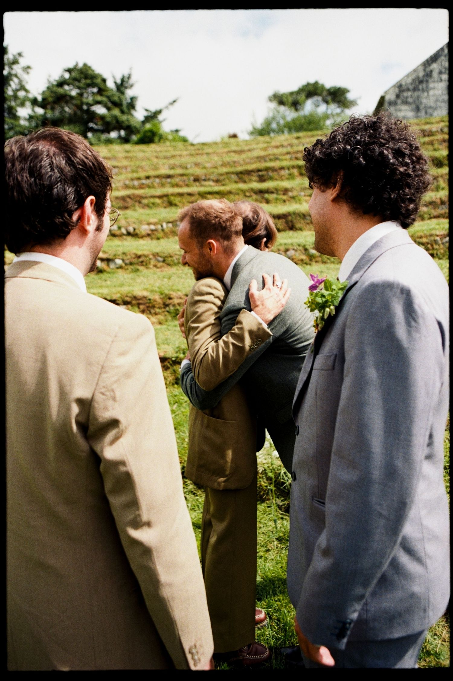 Groomsmen in suits sharing an emotional embrace at an outdoor wedding ceremony.