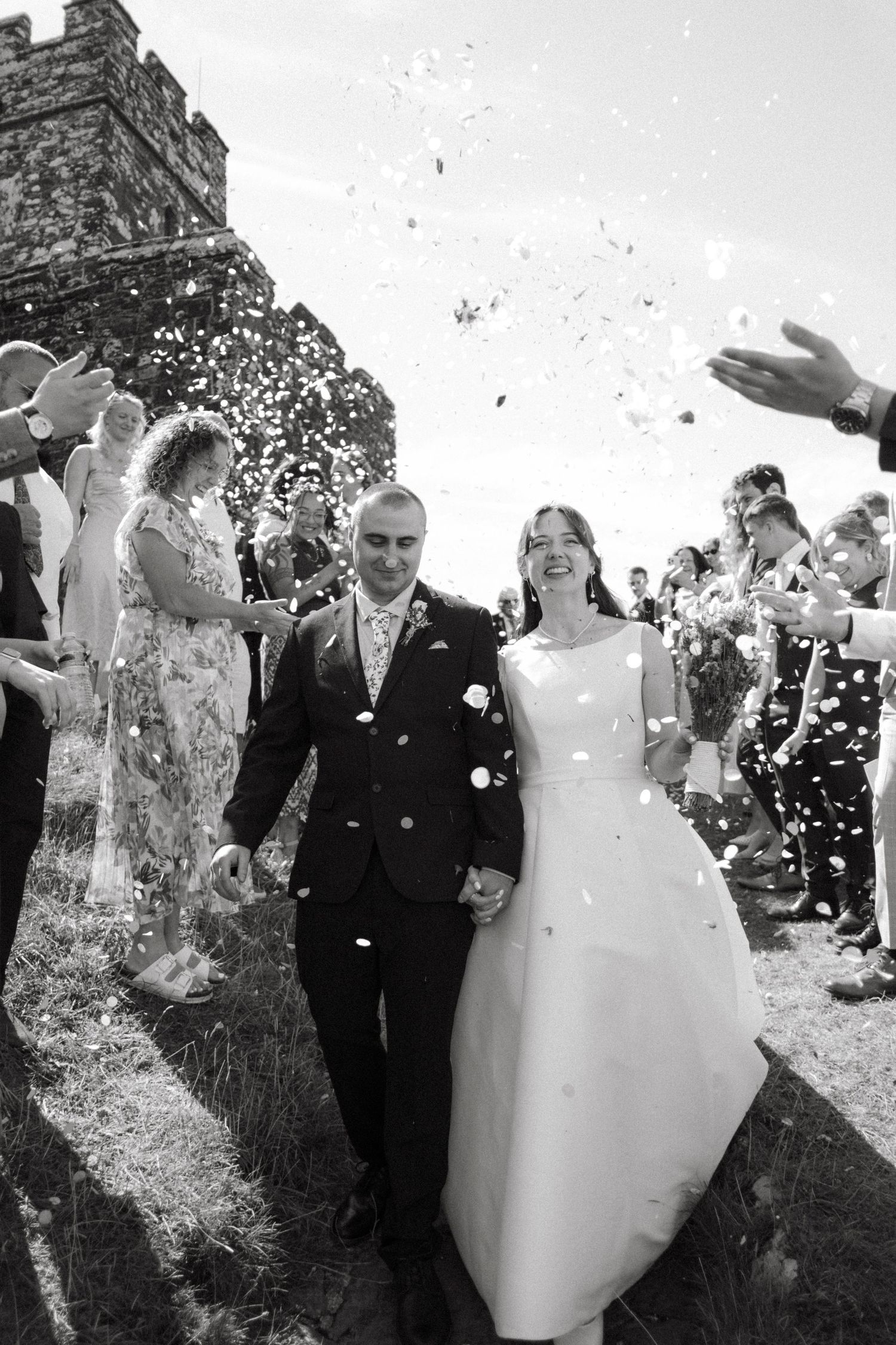 Newlyweds walking through a joyful confetti shower surrounded by cheering guests, black and white photo.