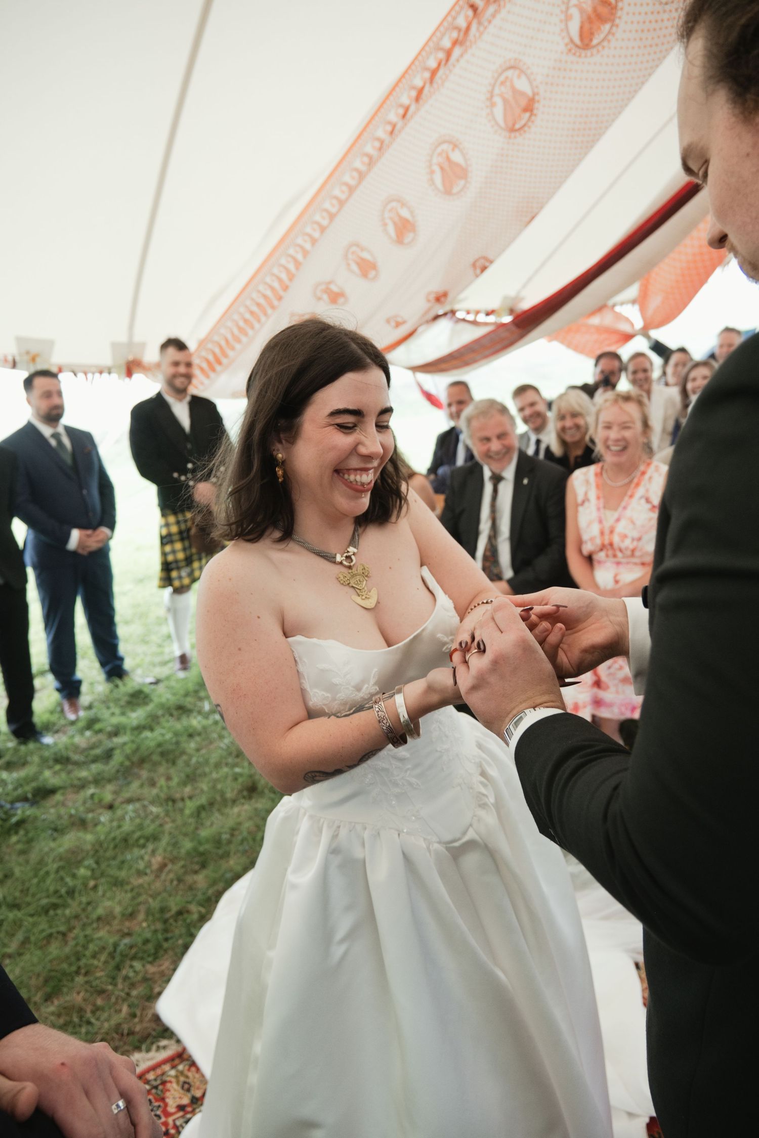Smiling bride in white strapless gown exchanges rings with groom under decorated tent at outdoor wedding ceremony.