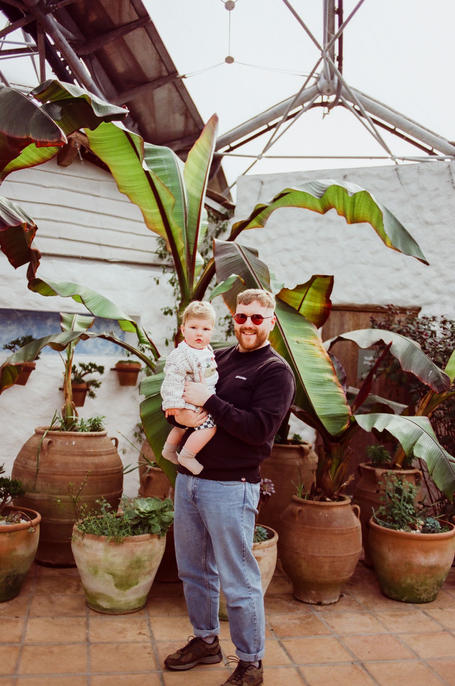 Red-bearded father in sunglasses holding his toddler among large potted banana plants in a garden.