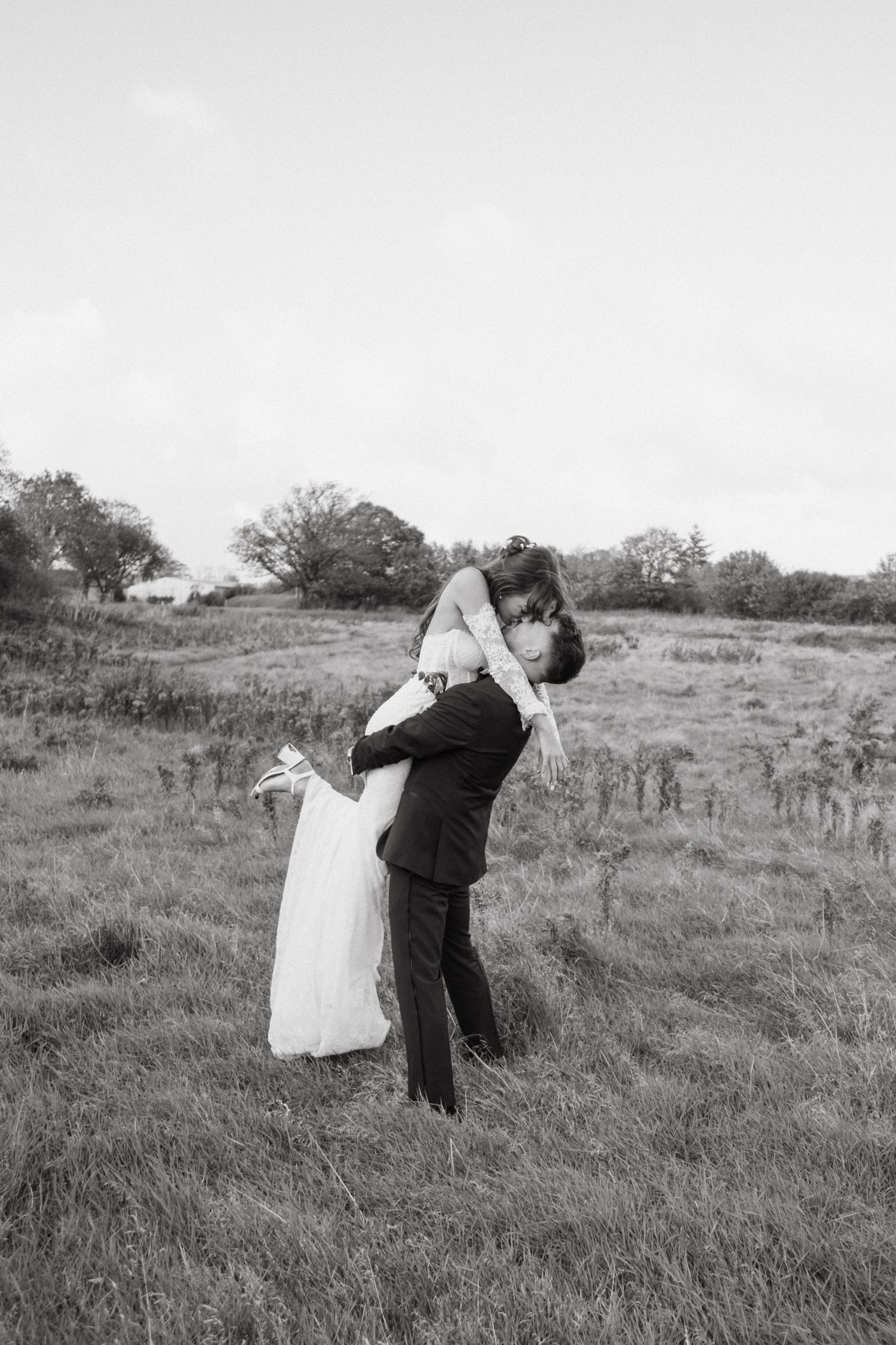 Groom lifts bride in white wedding dress in a romantic black and white photo taken in an open field.