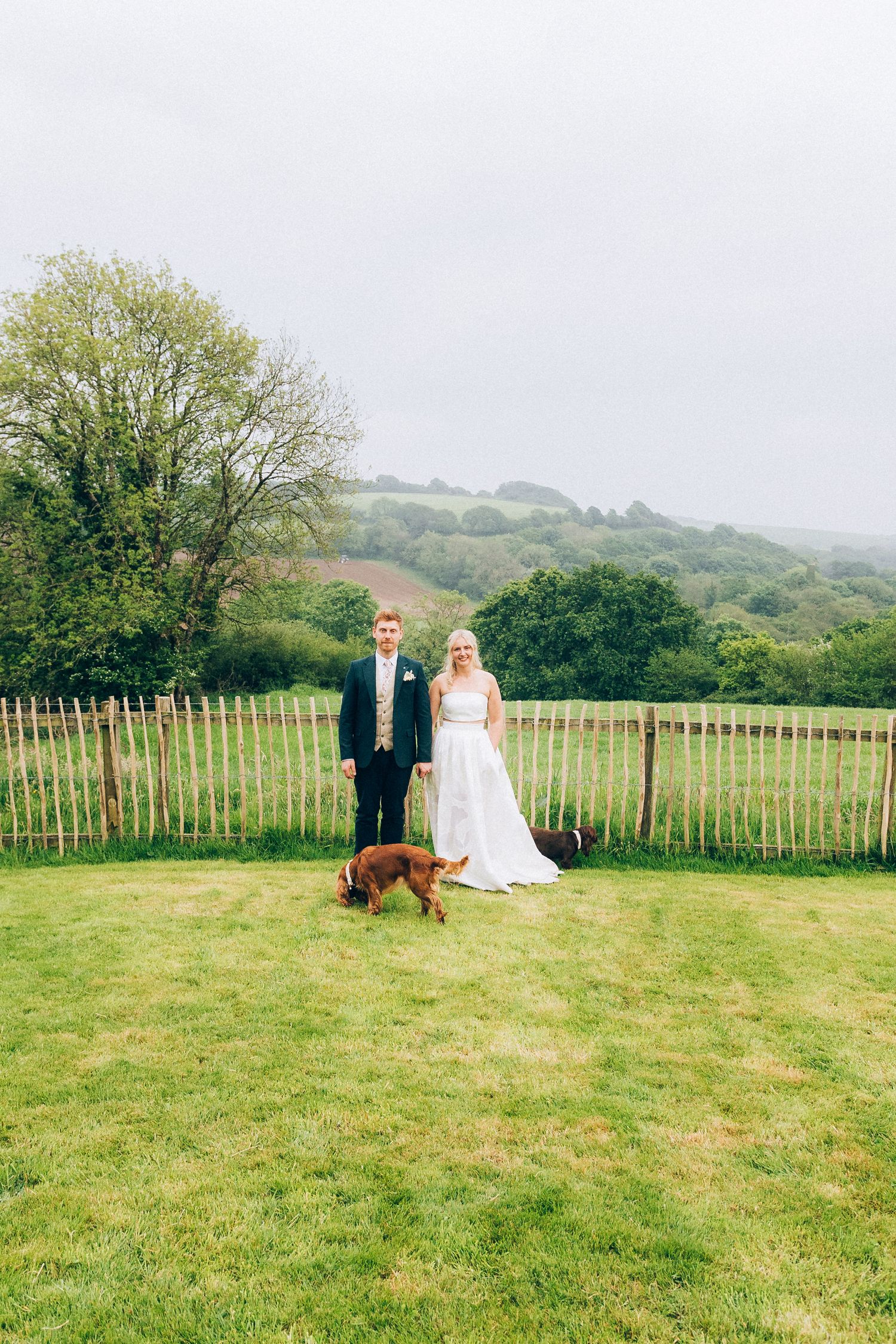 Bride and groom stand on a green hillside lawn with a brown dog and scenic countryside views behind them.