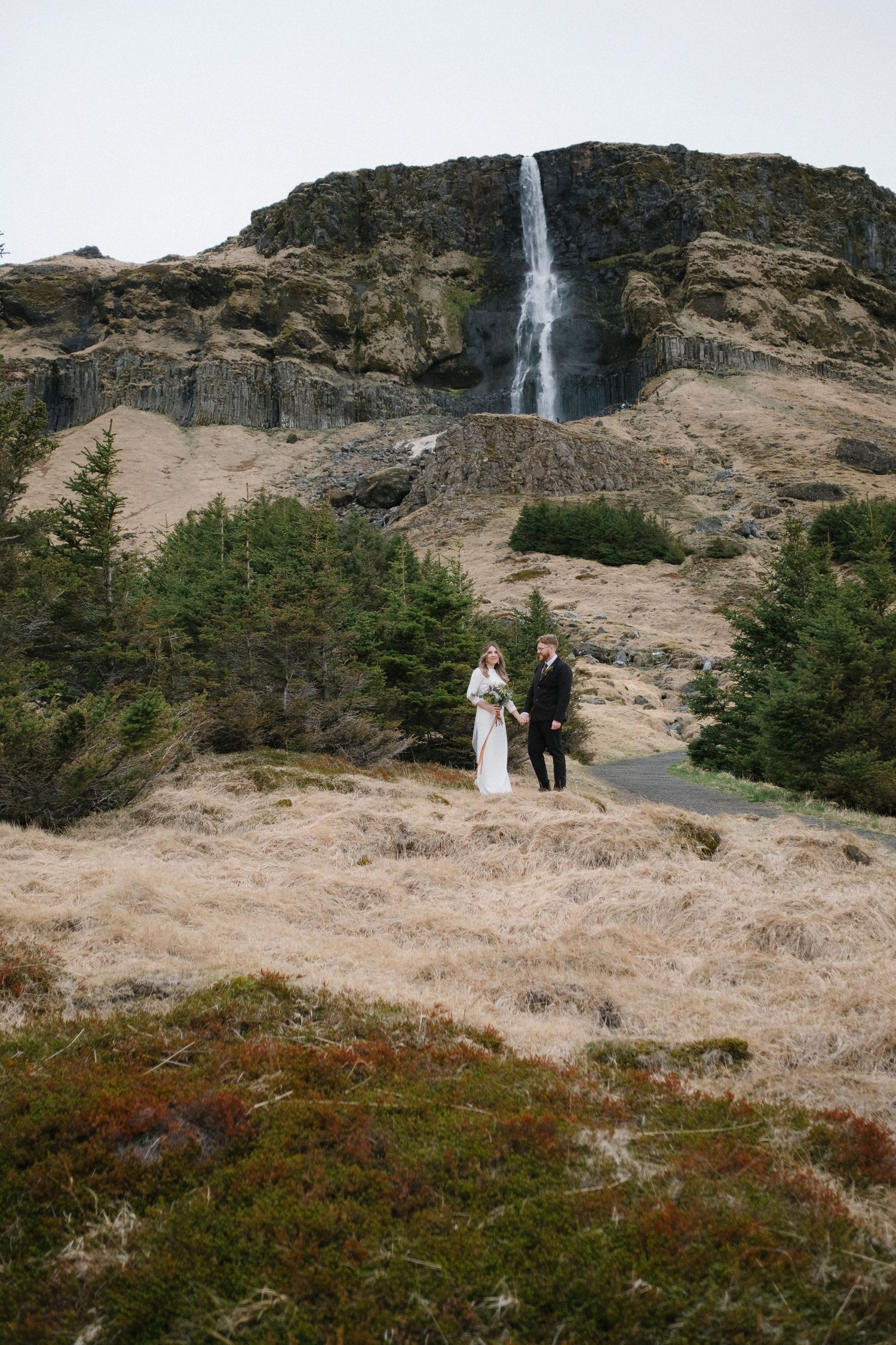 Bride and groom holding hands in front of a dramatic waterfall landscape.