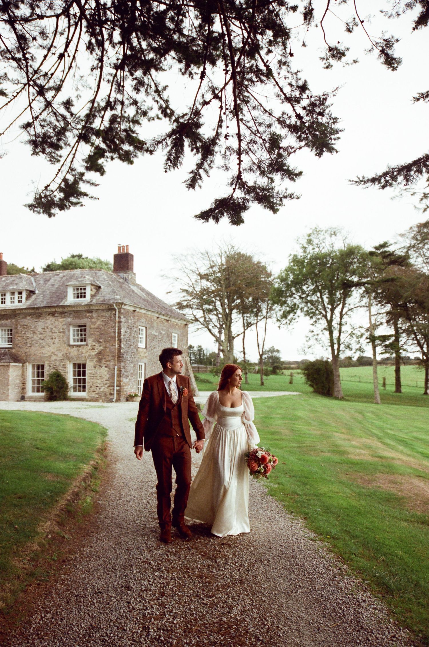 Bride and groom walk hand-in-hand on a gravel path outside a stone country house venue surrounded by lush greenery.