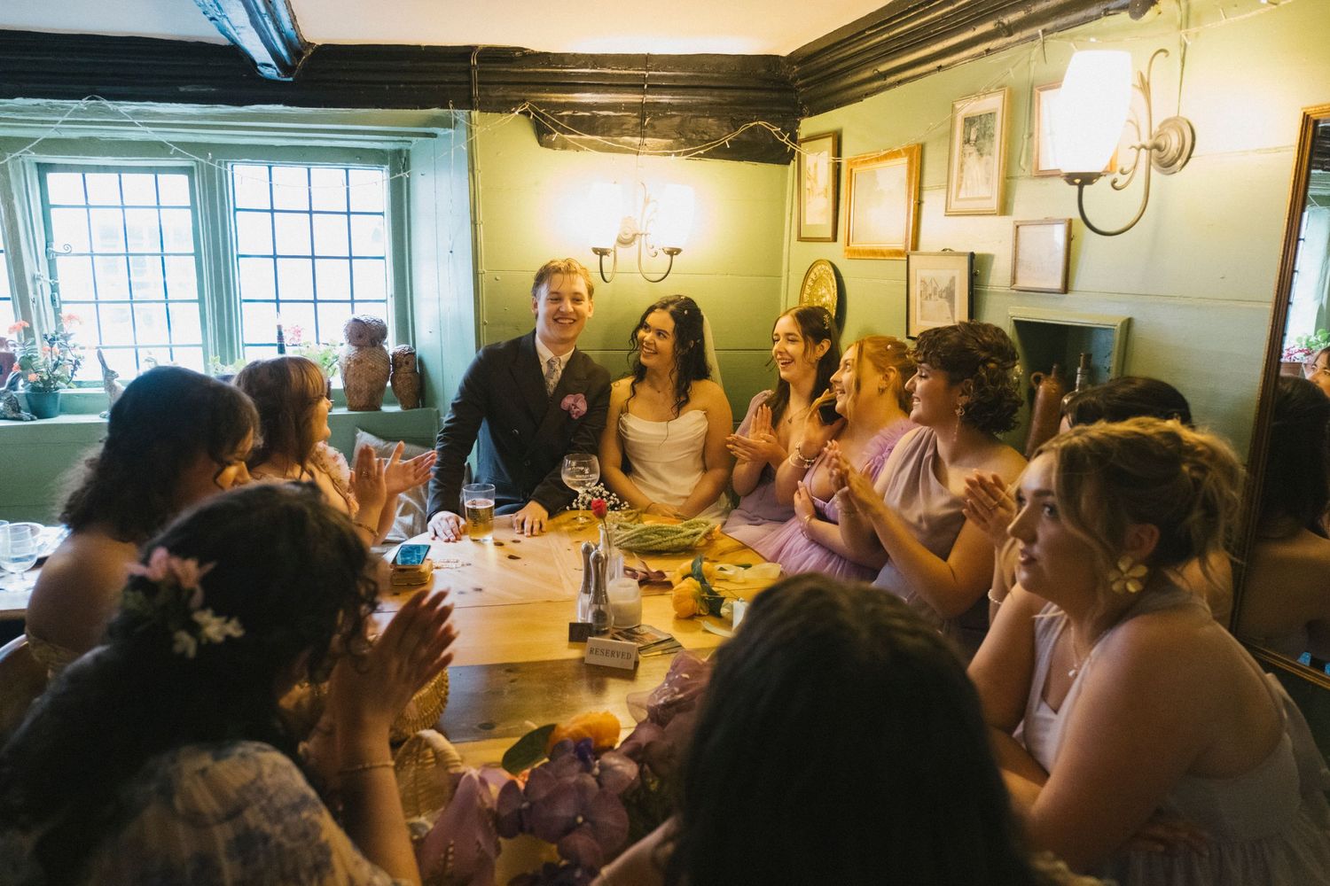 Bride and groom laughing with bridesmaids in lavender dresses at rustic wedding reception table.