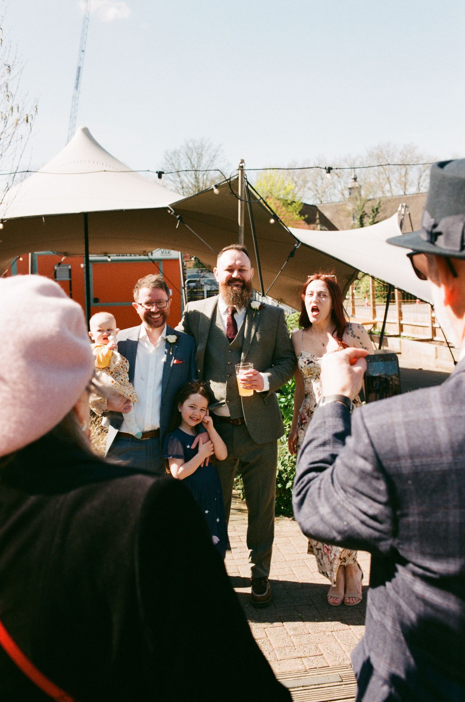 Guests laughing and celebrating outdoors at a wedding reception with a marquee tent in the background on a sunny day.