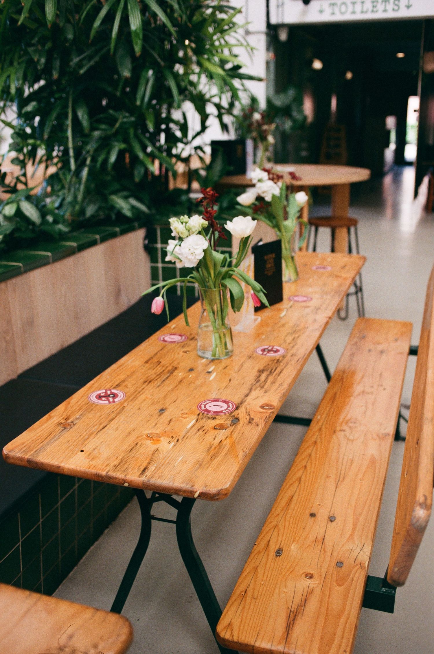 Rustic wooden picnic table with fresh flower centerpieces in a cozy green plant-filled cafe interior.