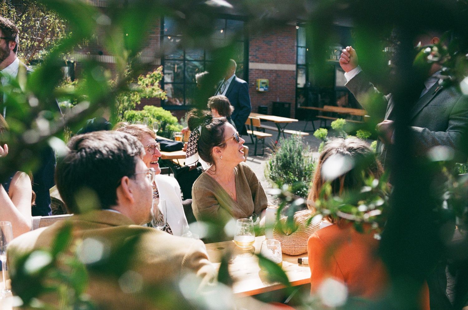 People dining outdoors at a sunlit restaurant patio surrounded by lush green plants on a bright day.