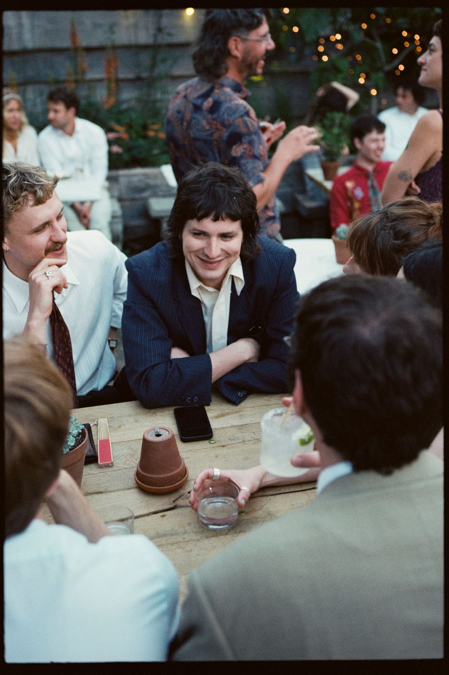 Guests socializing at an outdoor wedding reception party, seated around a wooden table with drinks and string lights.