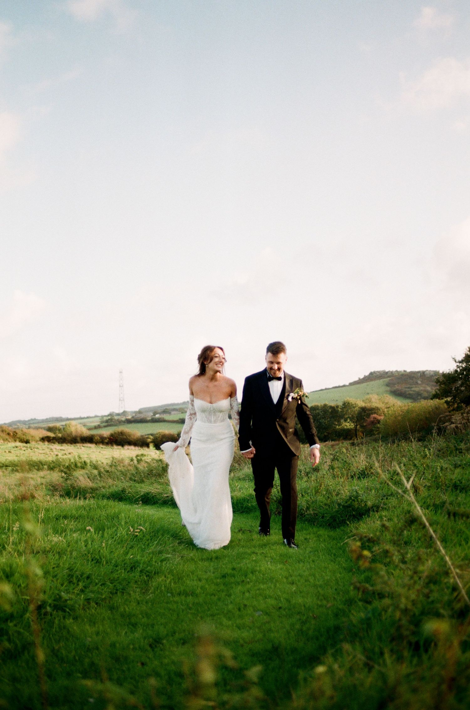 Bride in off-shoulder gown and groom in black tuxedo walking through green countryside fields laughing.