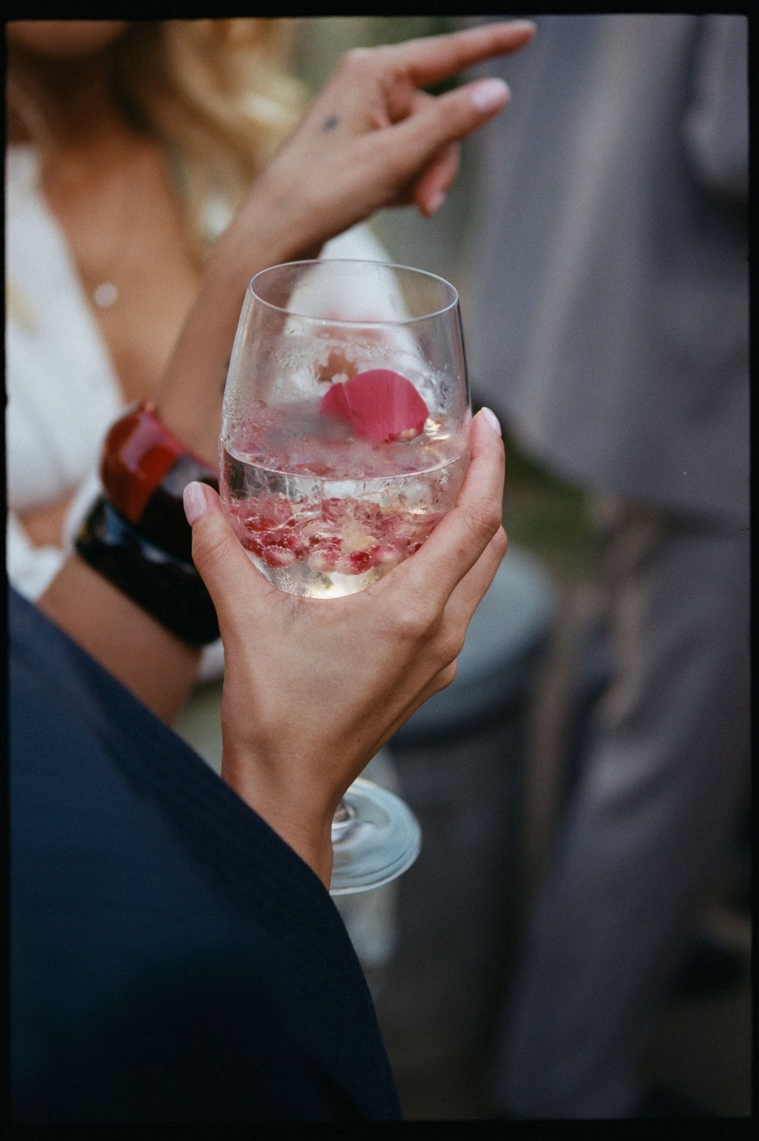 Close-up of hands holding a stemless wine glass with pink floral ice sphere at an outdoor wedding reception.