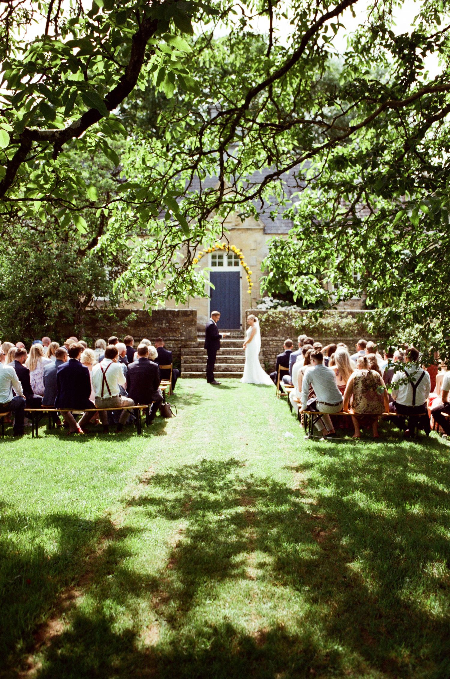 Outdoor wedding ceremony on lush green lawn with guests seated in rows beneath tree canopy leading to couple at altar.