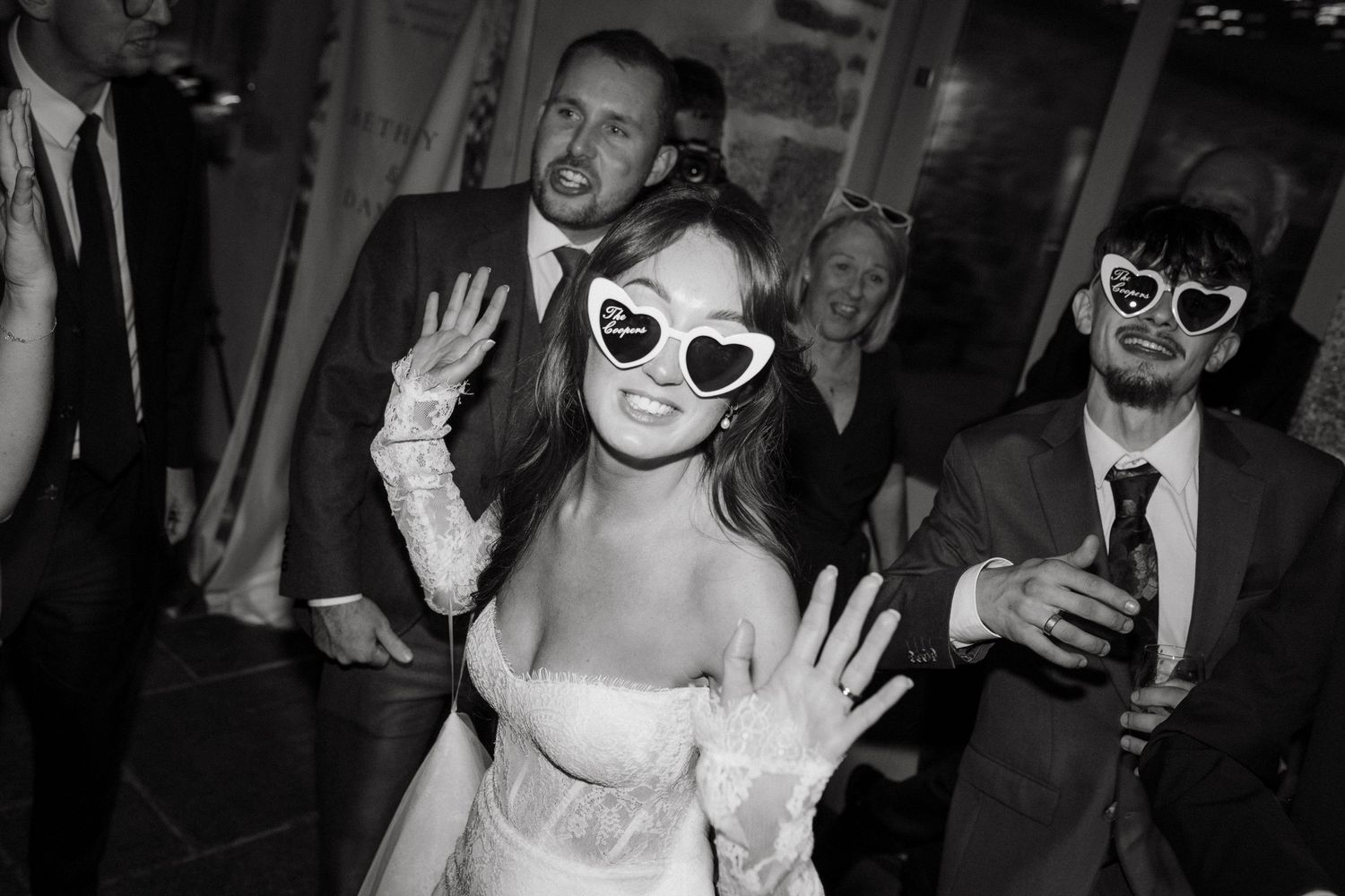 Bride wearing heart-shaped sunglasses dances joyfully with guests at black and white wedding reception.