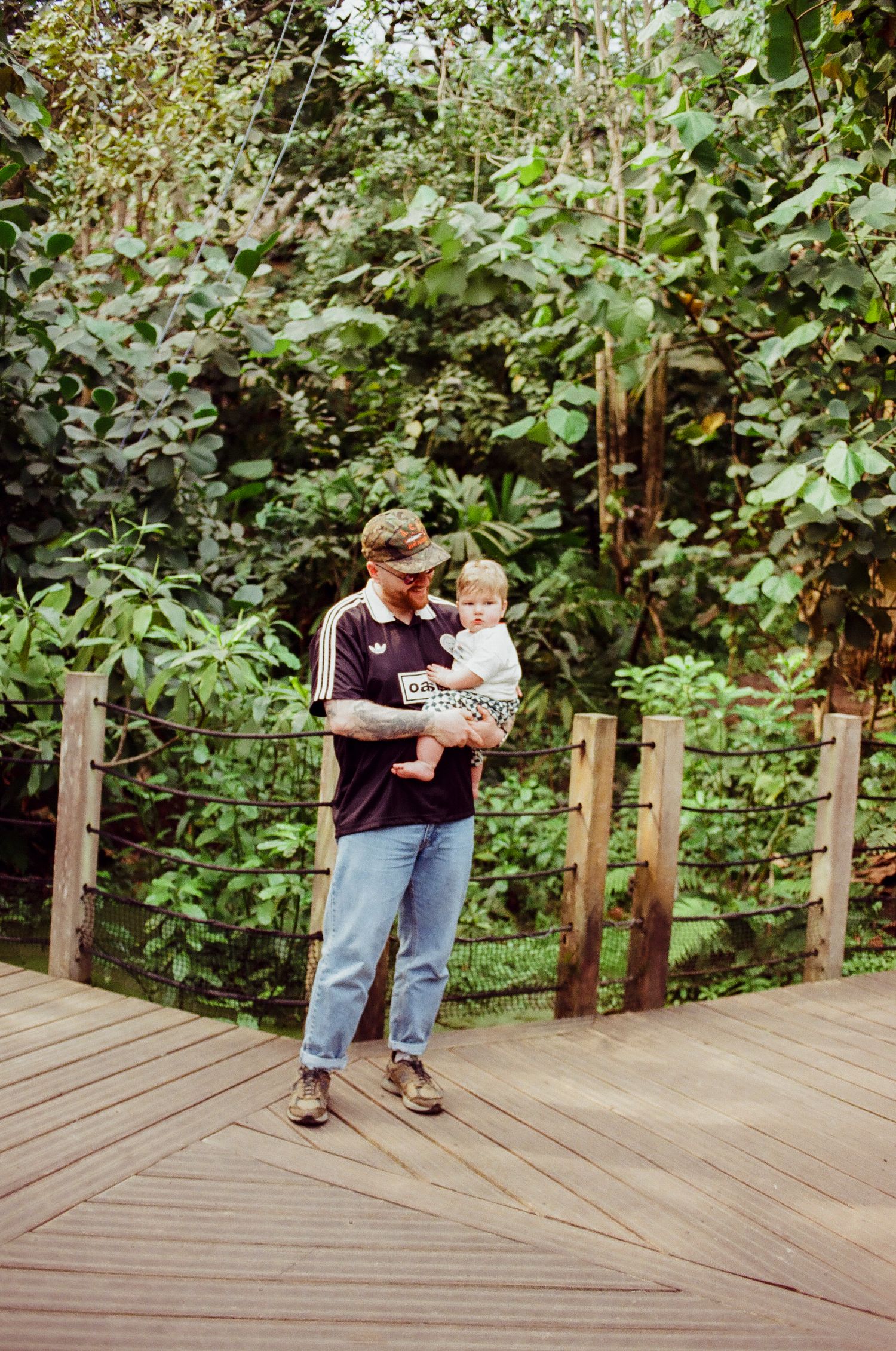 Father holding his baby on a wooden boardwalk surrounded by lush tropical greenery.
