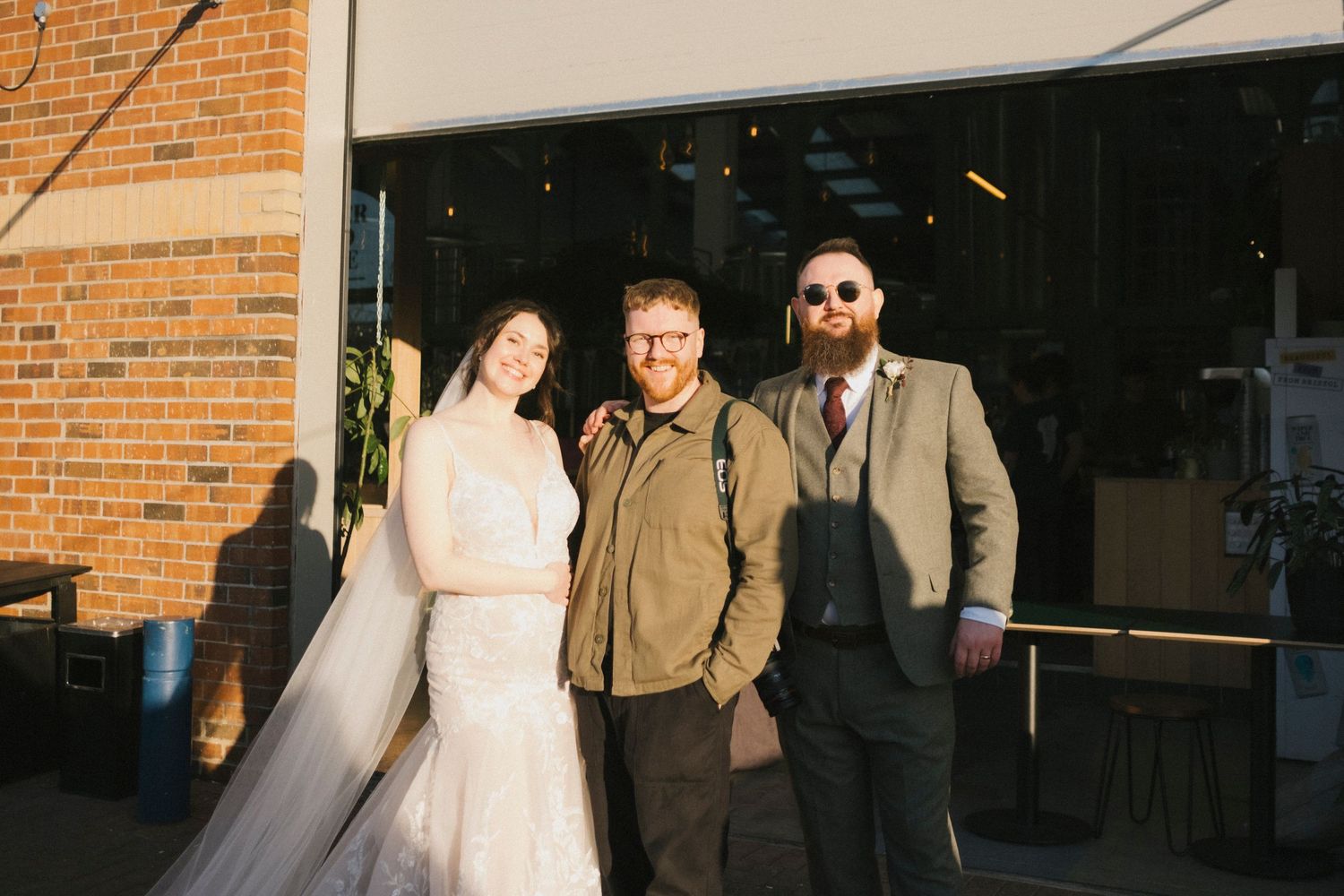 A bride in a white gown poses with two men outside a brick building at a wedding.