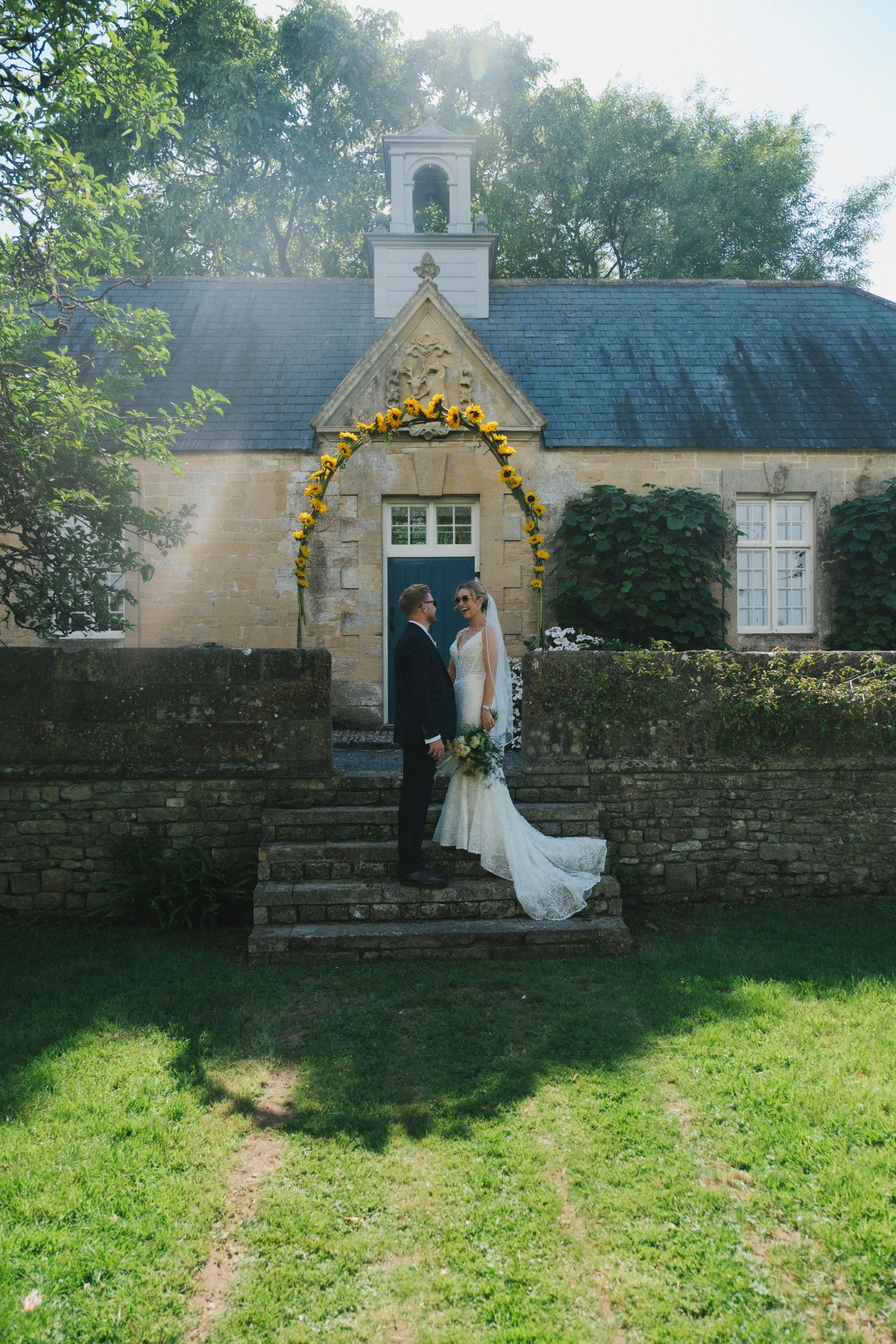 Bride and groom stand on stone steps before a charming chapel adorned with a yellow floral arch on their wedding day.