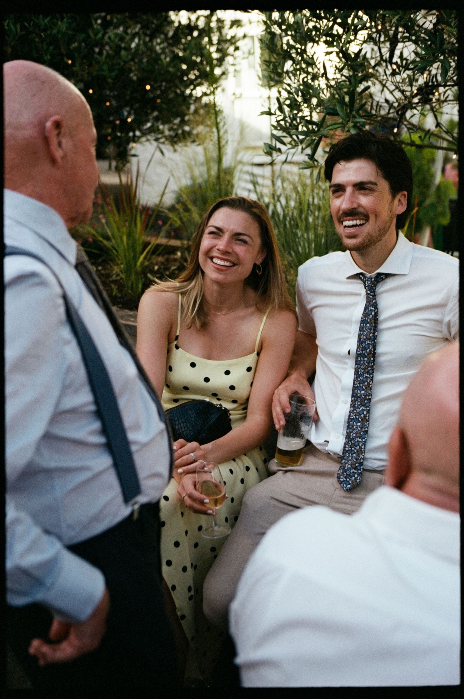 Happy couple laughing and enjoying drinks at an outdoor wedding reception surrounded by lush greenery.