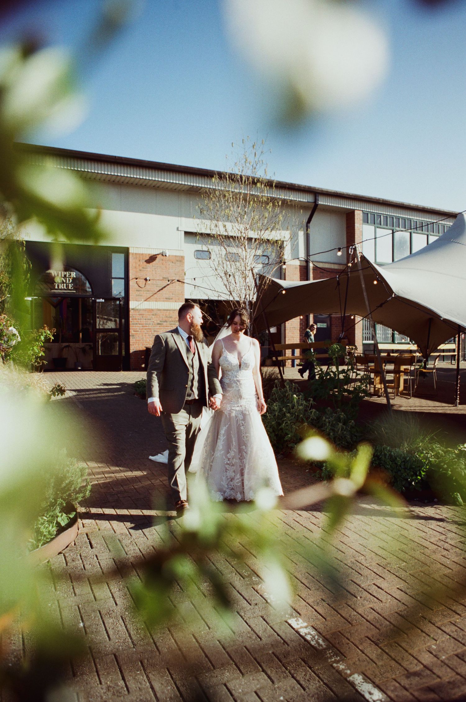 Bride in lace gown and groom in grey suit walk hand in hand outside a modern wedding venue on a sunny day.