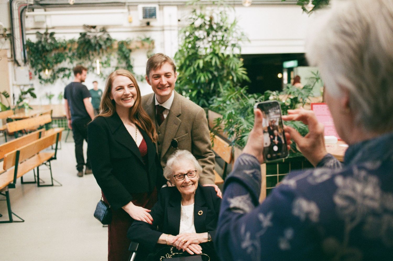 Two adults and an elderly woman pose for a photo indoors, surrounded by lush green plants in a bright venue.