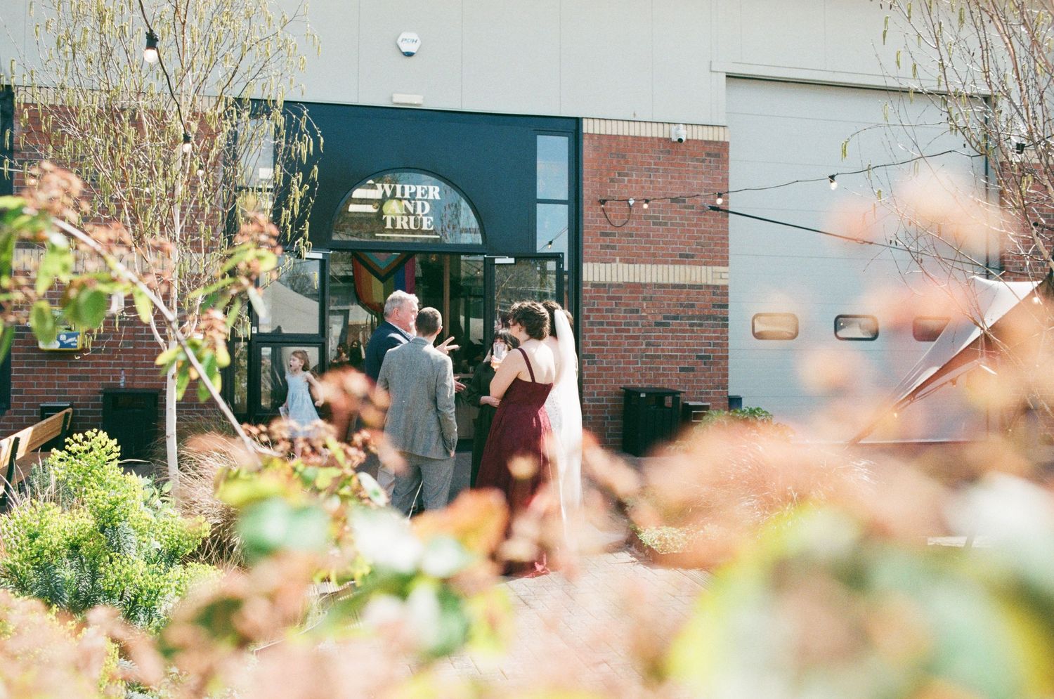 Couple in formal attire entering a brick building venue surrounded by lush greenery on a sunny day.