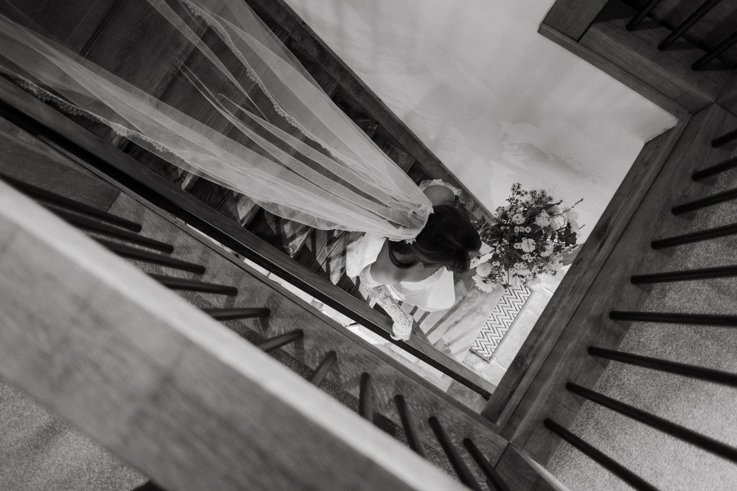 Aerial black and white view of a bride descending a wooden staircase holding a bouquet on her wedding day.