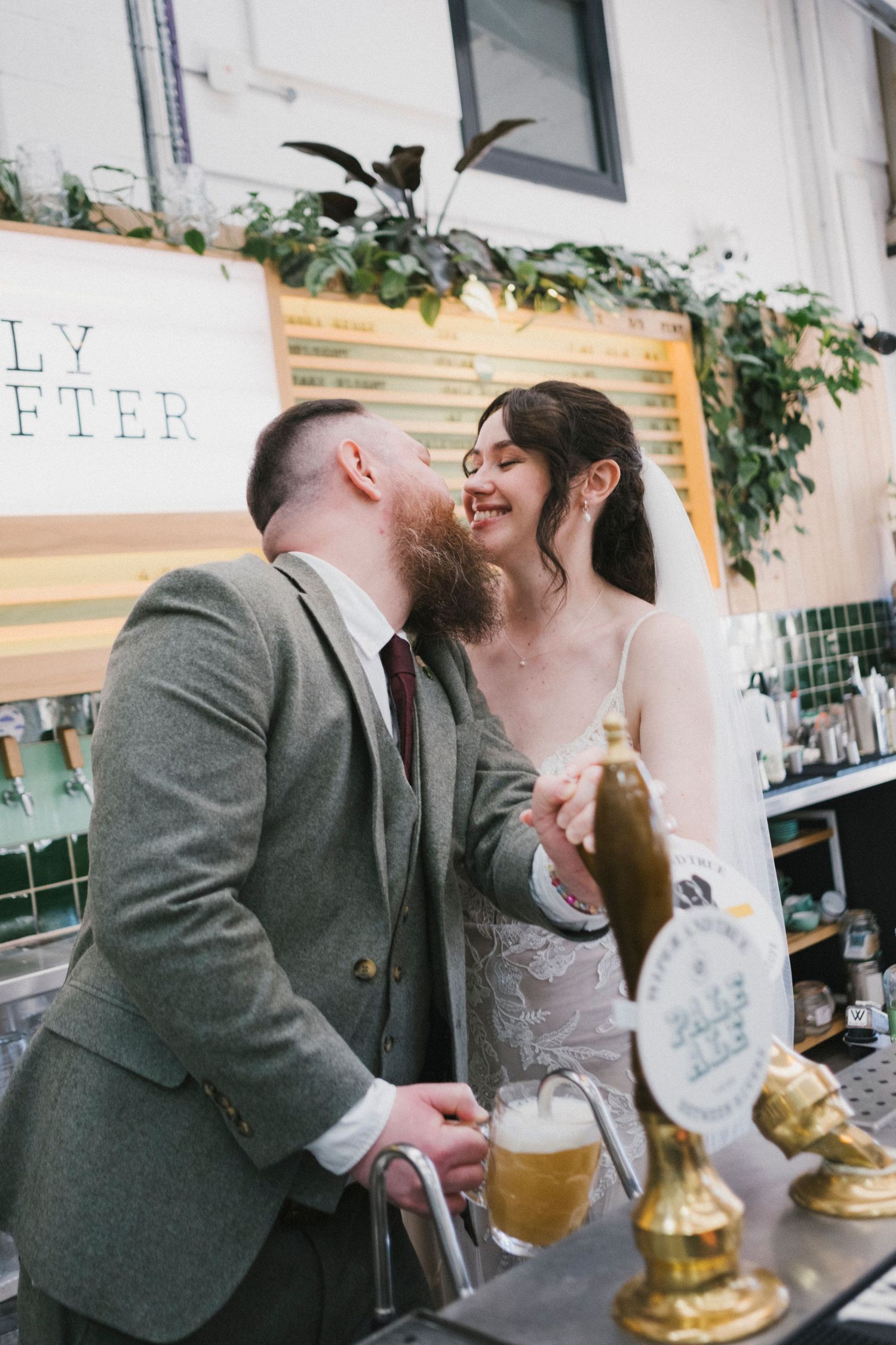 Bride and bearded groom sharing a kiss at a bar with beer taps at their wedding reception.