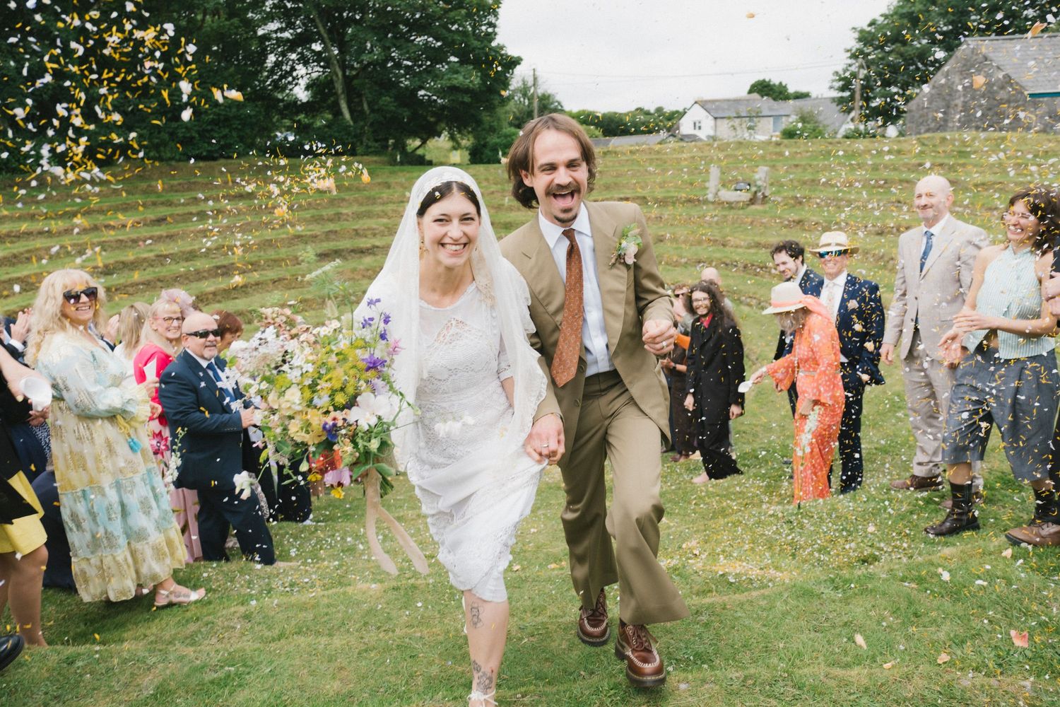 Joyful newlyweds walk through confetti shower surrounded by cheering guests at an outdoor countryside wedding celebration.