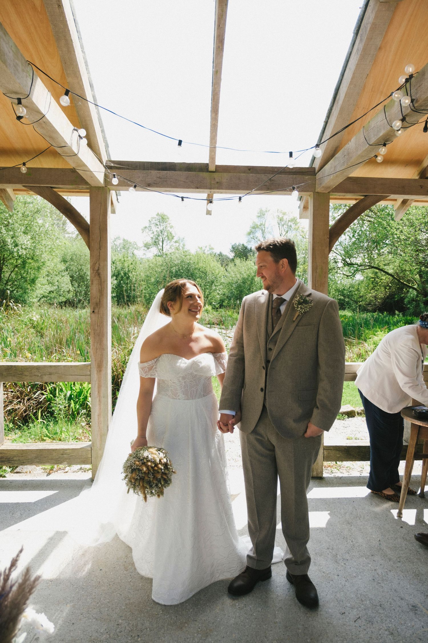 Bride and groom holding hands smiling at each other inside a sunlit wooden barn wedding venue.