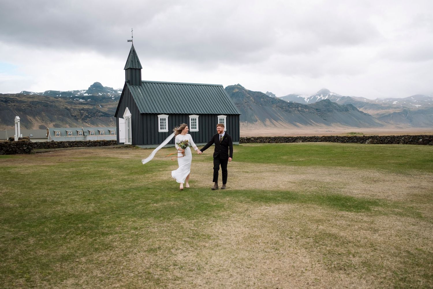 A bride and groom walk hand in hand on a green field beside a black church in Iceland.