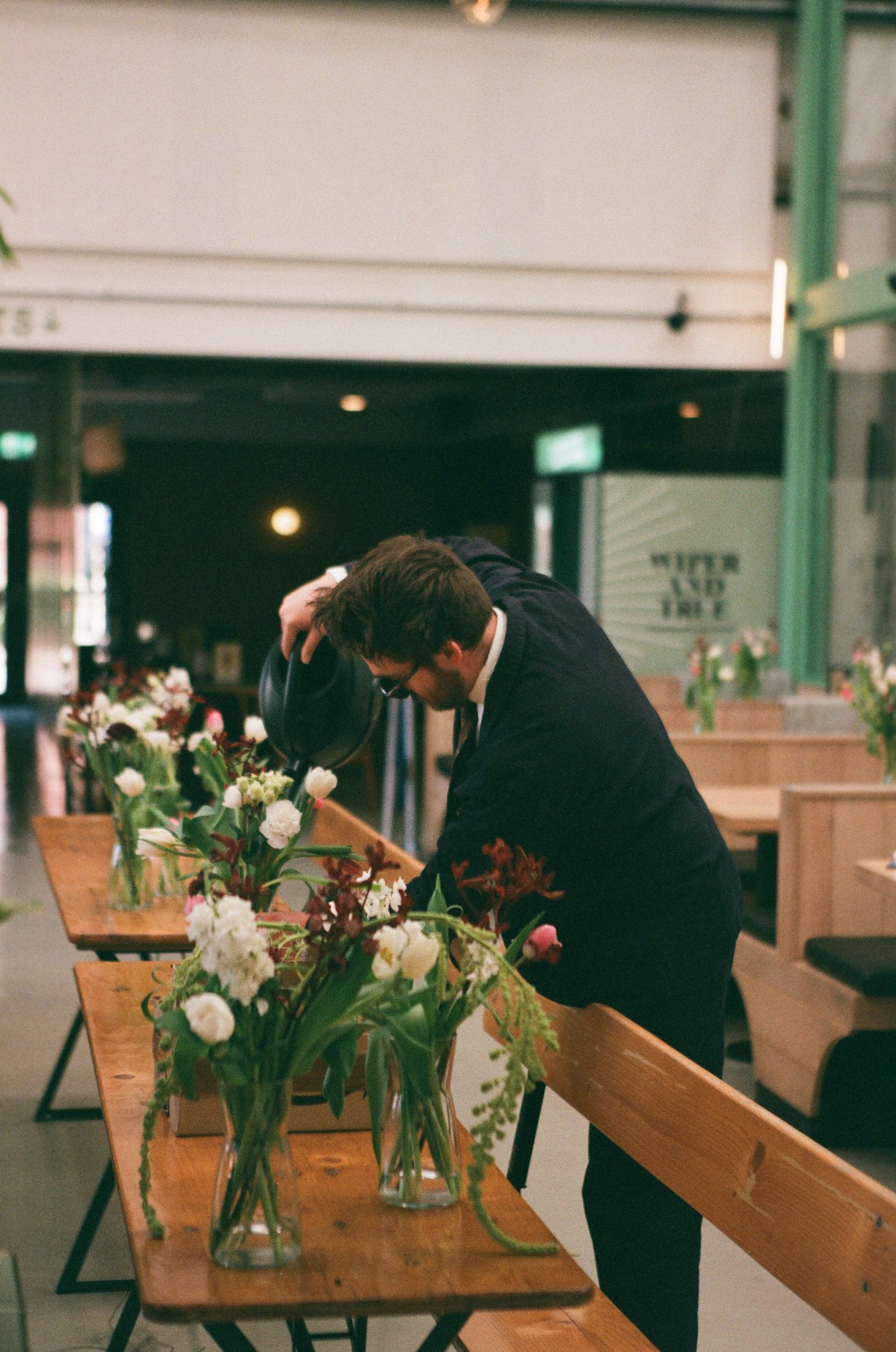 Man in black suit arranging fresh flowers in glass vases on wooden tables in an elegant indoor venue.
