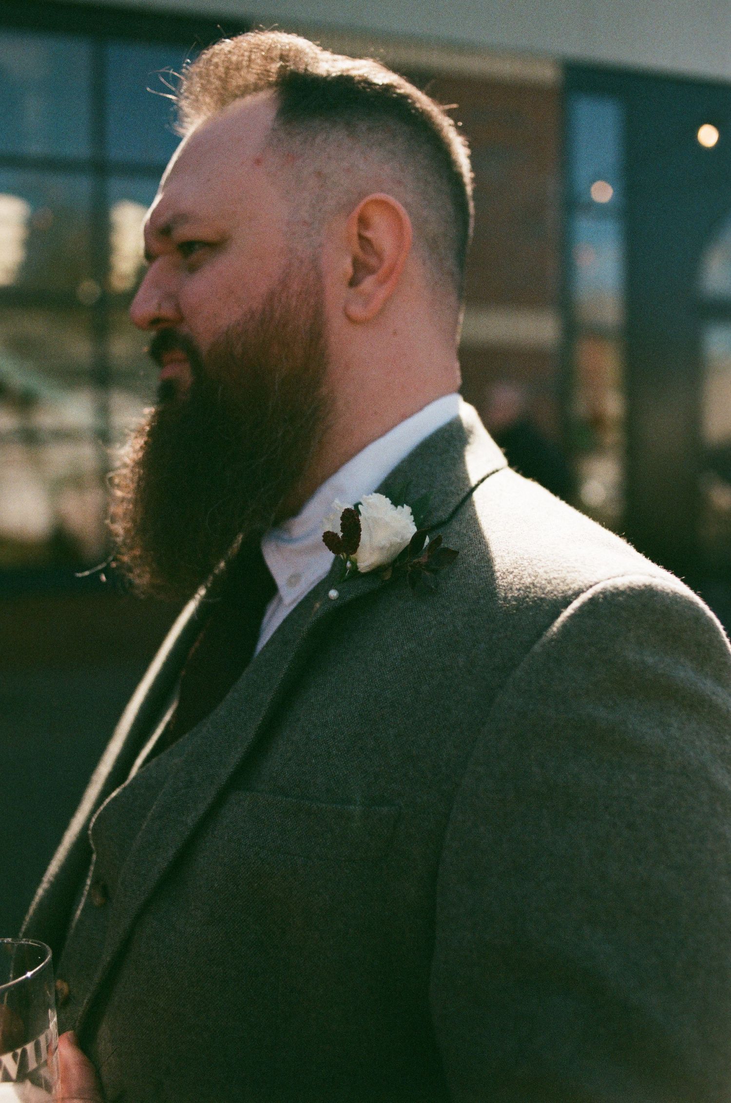 Bearded man in gray suit with white boutonniere, profile view, at outdoor wedding event.
