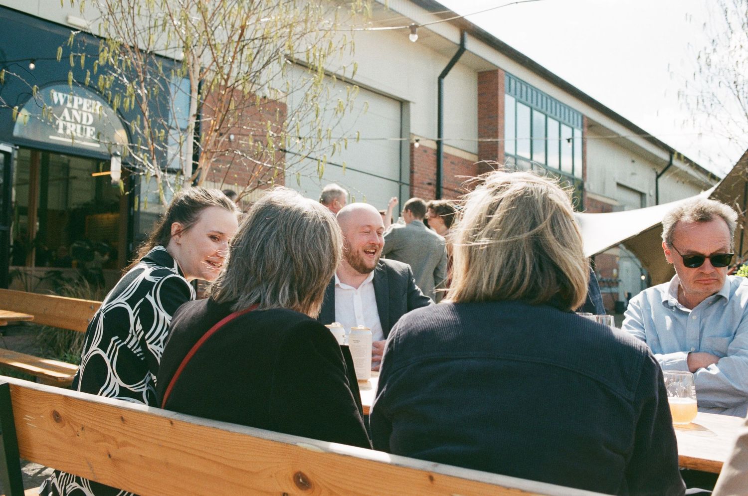 Group of friends laughing and socializing at an outdoor cafe patio on a sunny day.