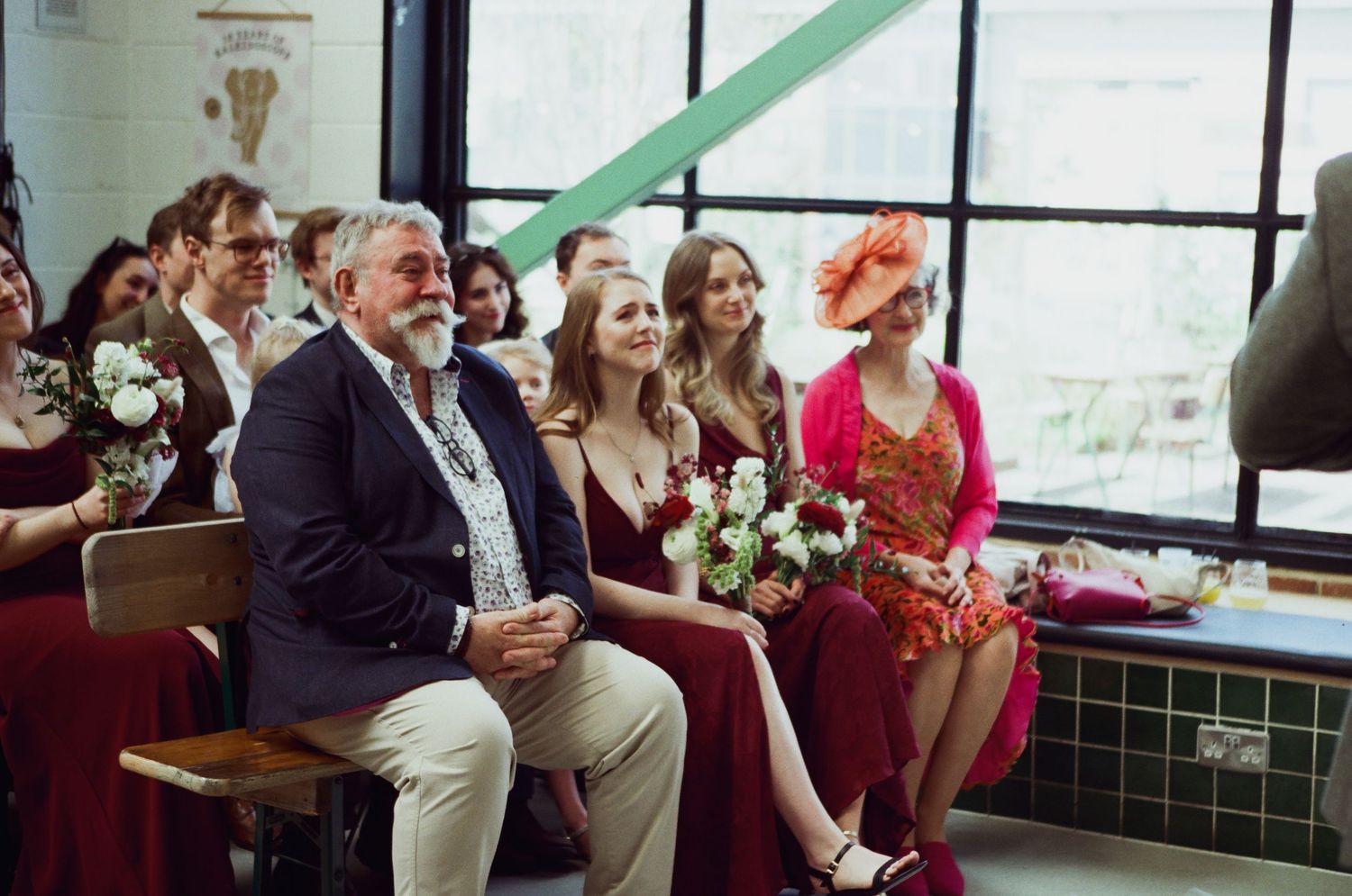 Wedding guests seated on wooden benches, including bridesmaids holding bouquets, smiling during a ceremony.