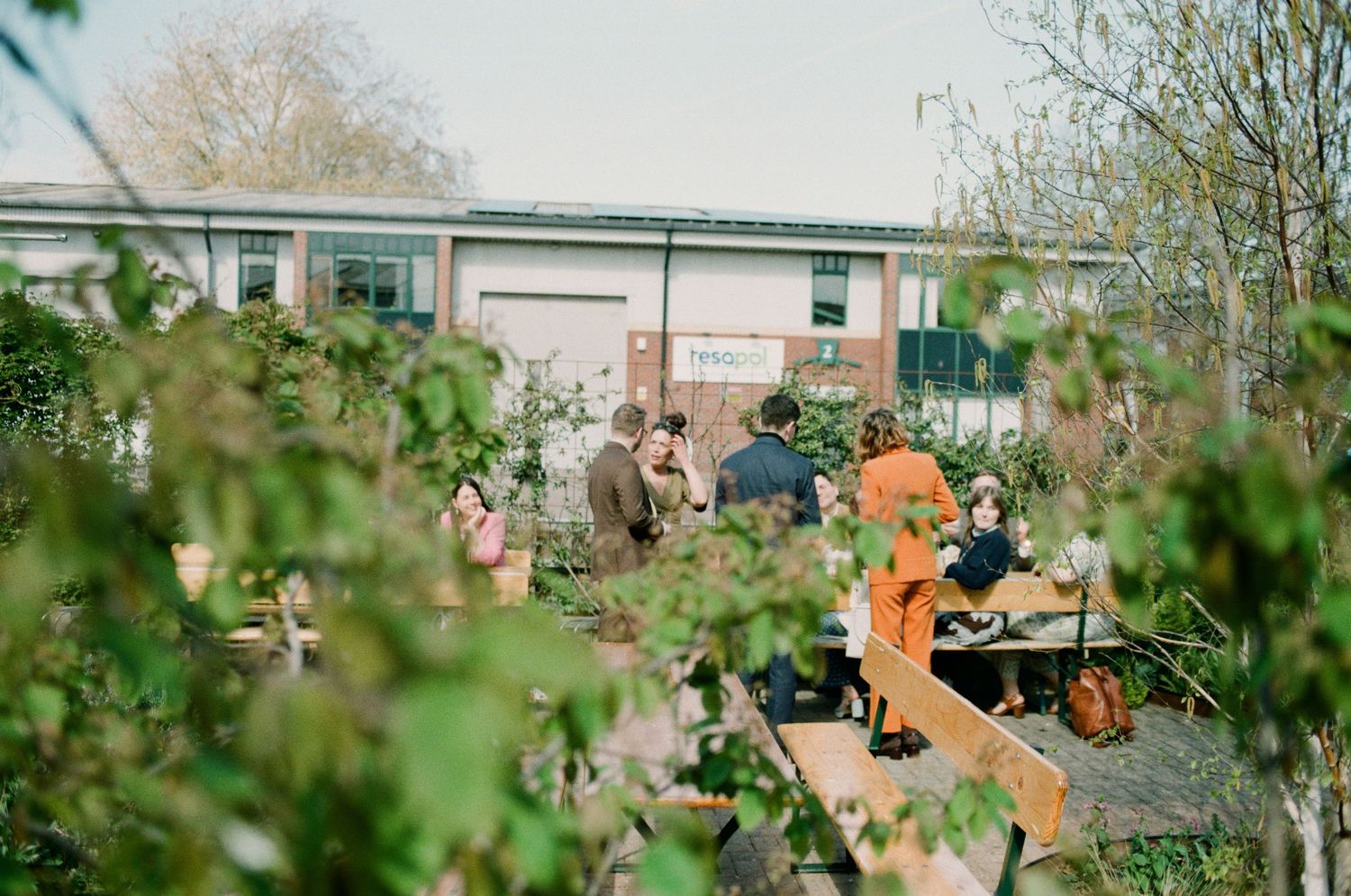 People gather in a lush community garden surrounded by green plants near an industrial building on a bright spring day.