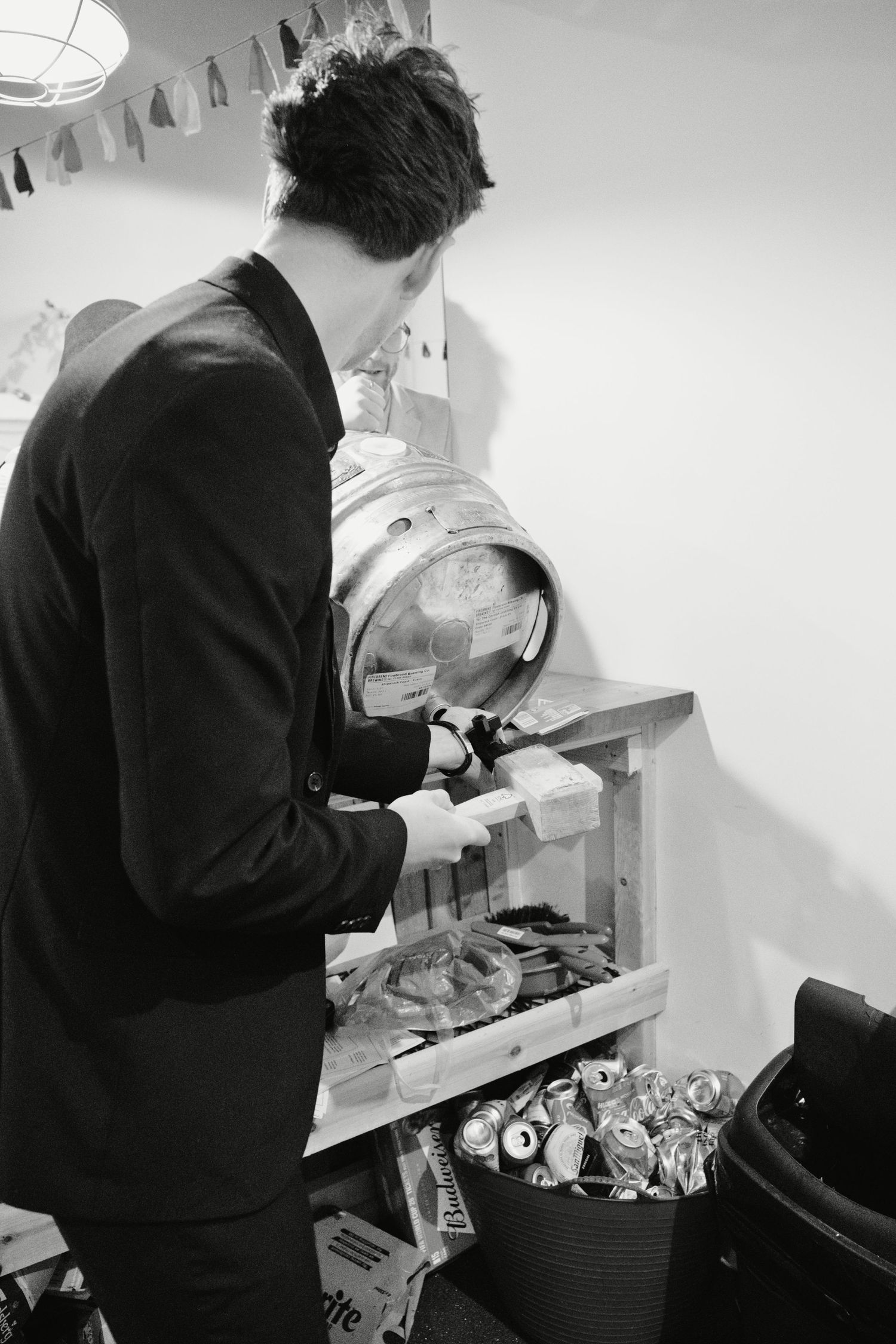 Man in black suit tapping a beer keg on a wooden stand at a party event, black and white photo.